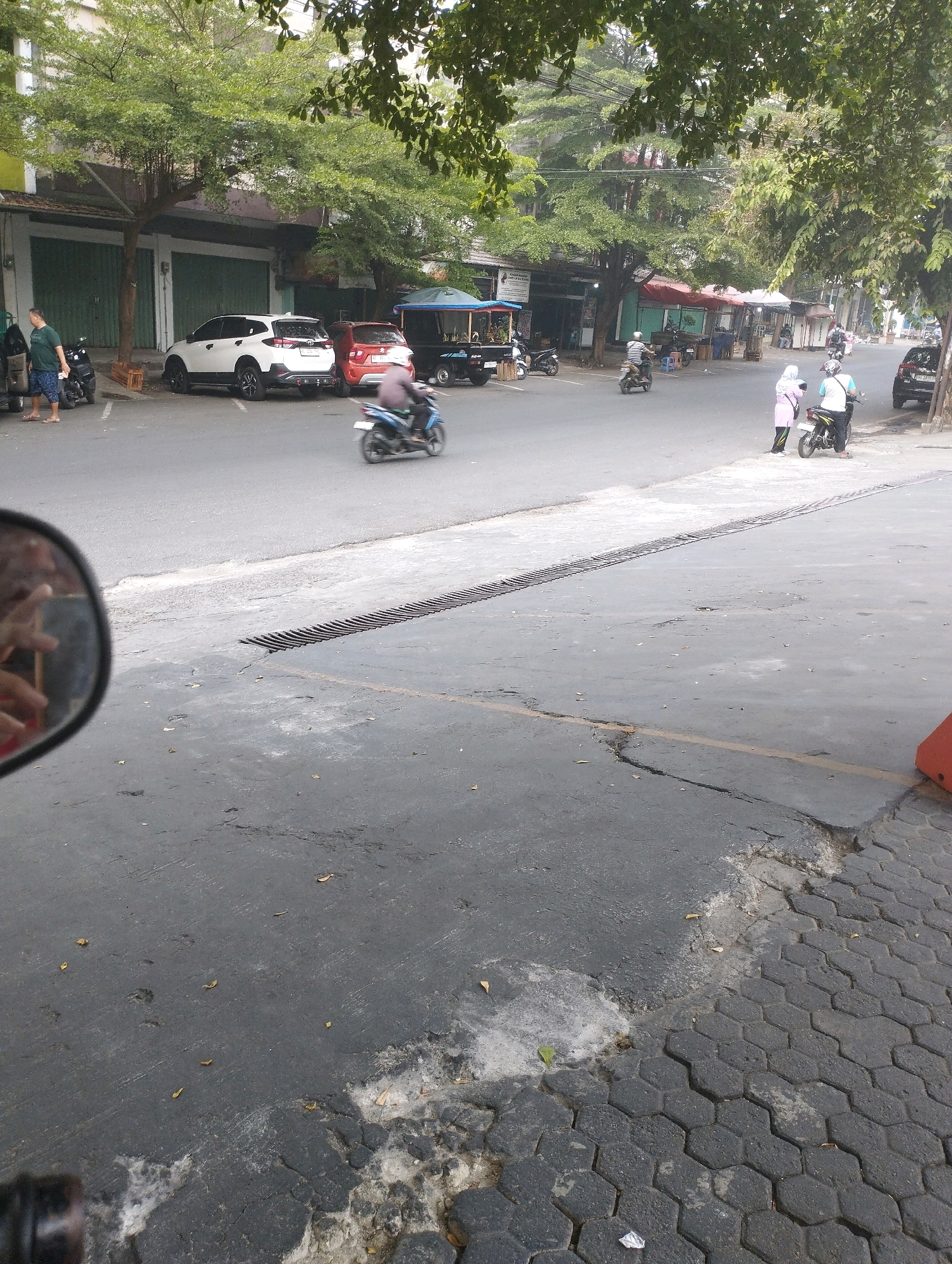 The image captures a street scene in Talang Jawa, Indonesia, with a focus on the asphalt road and the surrounding environment. In the foreground, a motorcycle is seen in motion, indicated by motion blur, on the left side of the frame. A side mirror, likely from a vehicle, is visible in the lower left corner, reflecting what appears to be a hand. The road surface shows signs of wear and tear, with cracks and patches of lighter-colored material. A metal grate, possibly a drain cover, runs horizontally across the road.
In the mid-ground, several parked cars, including a white SUV and a red hatchback, are visible along the side of the street. A man in a green shirt and shorts stands near the parked cars. Further down the street, a blue canopy tent is set up, and several people are seen near shops or stalls. More motorcycles are parked and in motion. The background reveals a row of buildings with closed green metal doors, and lush green trees overhang the street. The overall impression is a typical urban street environment in Indonesia.