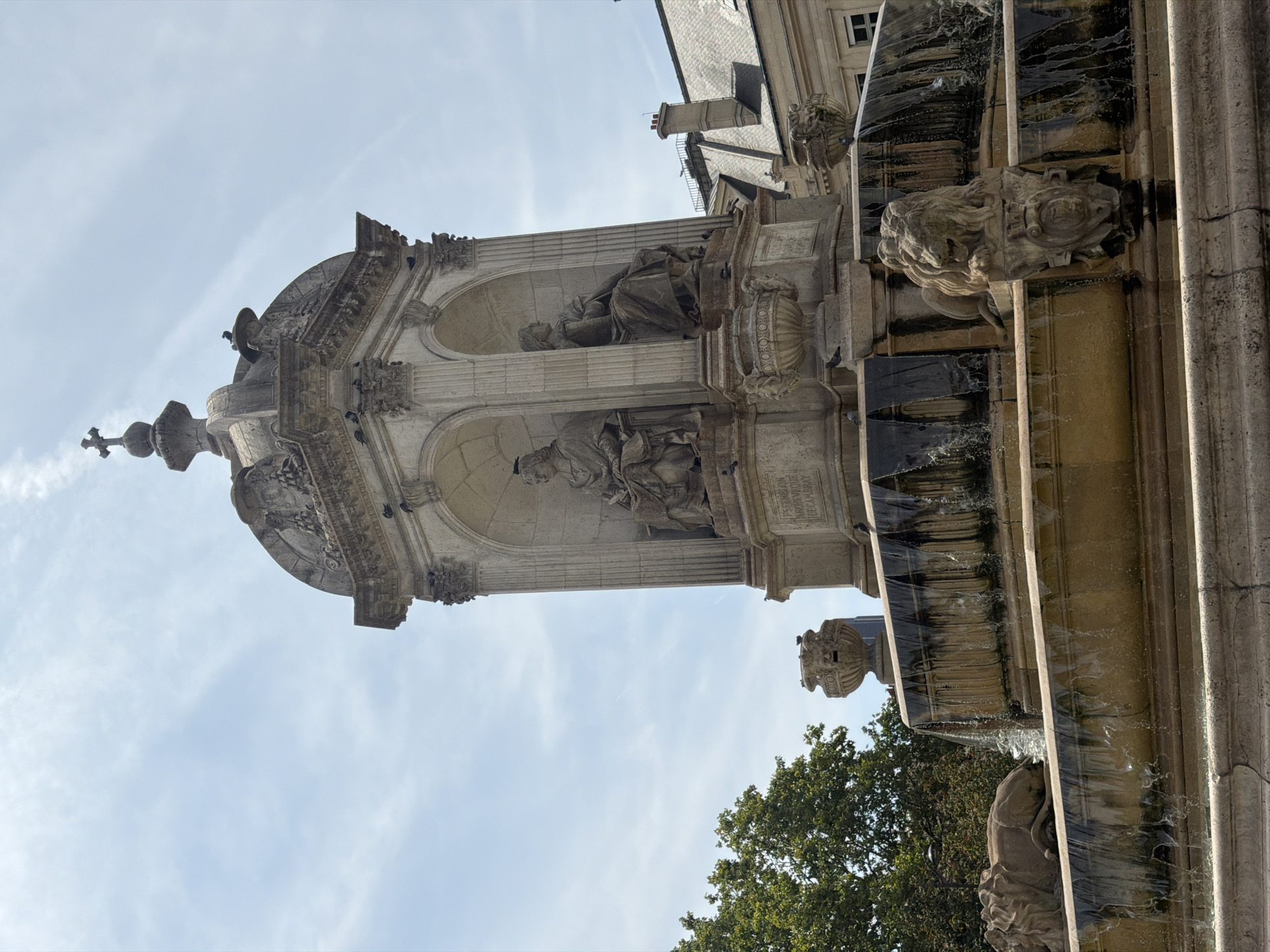 The image shows the Fontaine des Innocents in Paris, France. It is a 16th-century fountain that was later enlarged and embellished by sculptor Jean-Baptiste Belloc in the 19th century. The fountain is made of stone and features several sculptures, including four statues of nymphs and a central dome topped with a cross. The fountain is surrounded by a fountain basin with lion sculptures and flowing water. The background shows buildings and trees, indicating an urban environment. The sky is clear with some clouds.