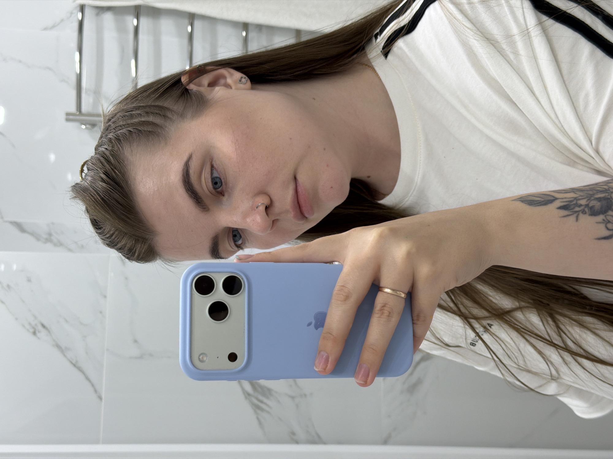 A woman with long brown hair is holding a blue iPhone in her right hand, taking a selfie in a bathroom. The background includes white marble tiles and a chrome towel rack. The woman is wearing a white t-shirt with black stripes on the shoulders. She has a nose ring in her left nostril and a tattoo behind her left ear and on her left forearm. The photograph appears to be taken in a bathroom with white marble tiles, which possibly indicates a residential or hotel setting. The city of Chaikovsky, Russia, cannot be confirmed as the location based solely on the visual information available in the image.
