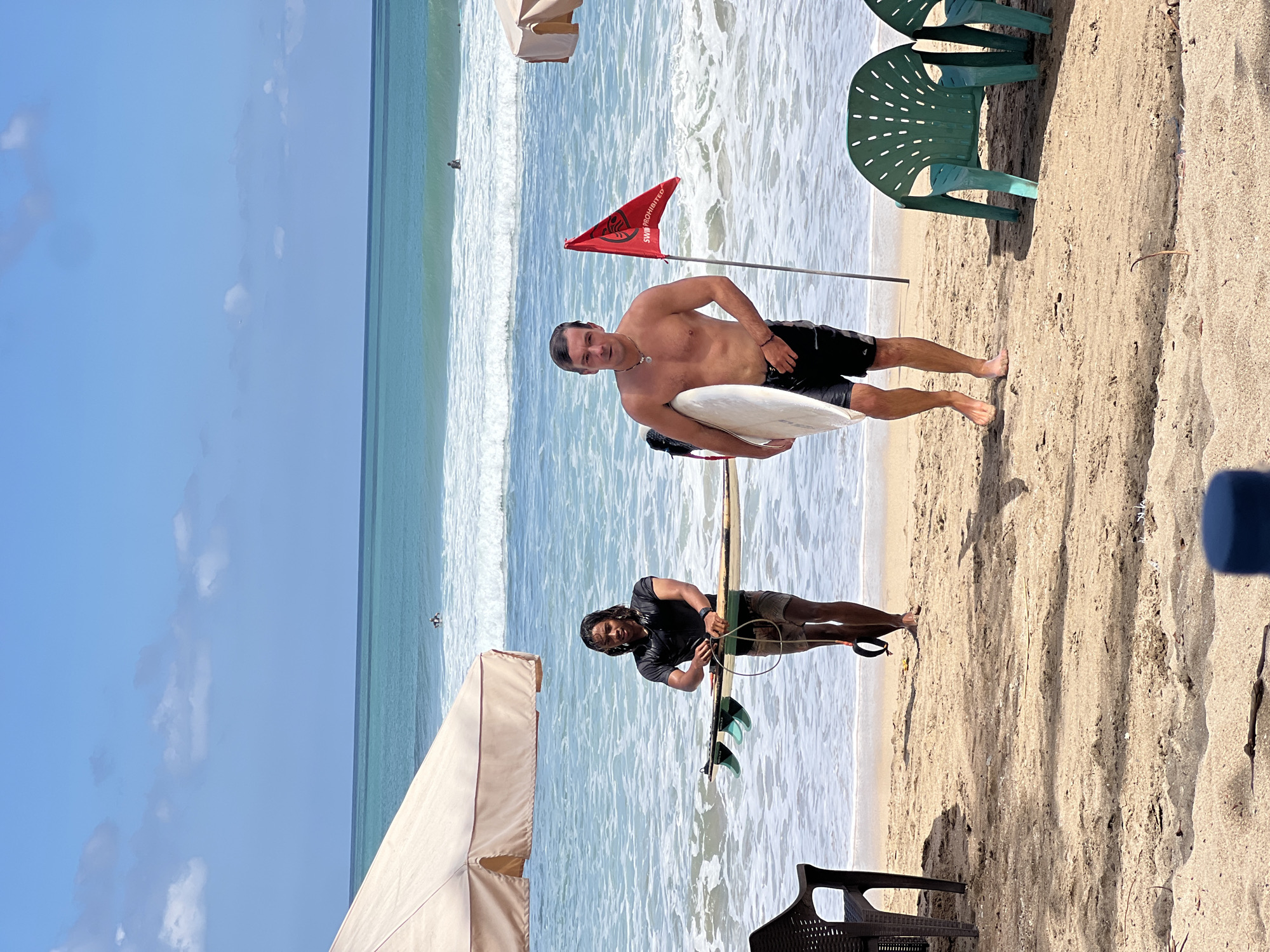 Two men are walking on a sandy beach in Kuta, Indonesia. The man on the left, with long dark hair and wearing a black t-shirt and shorts, carries a surfboard horizontally. The man on the right, with short brown hair and shirtless, carries a white surfboard under his arm. Both men are walking towards the viewer. A red flag with white text is planted in the sand behind the man on the right. The ocean with waves is visible in the background, and a few surfers are in the water. There are beach umbrellas and chairs on the sand, suggesting a popular tourist spot. The sky is blue with some scattered clouds.