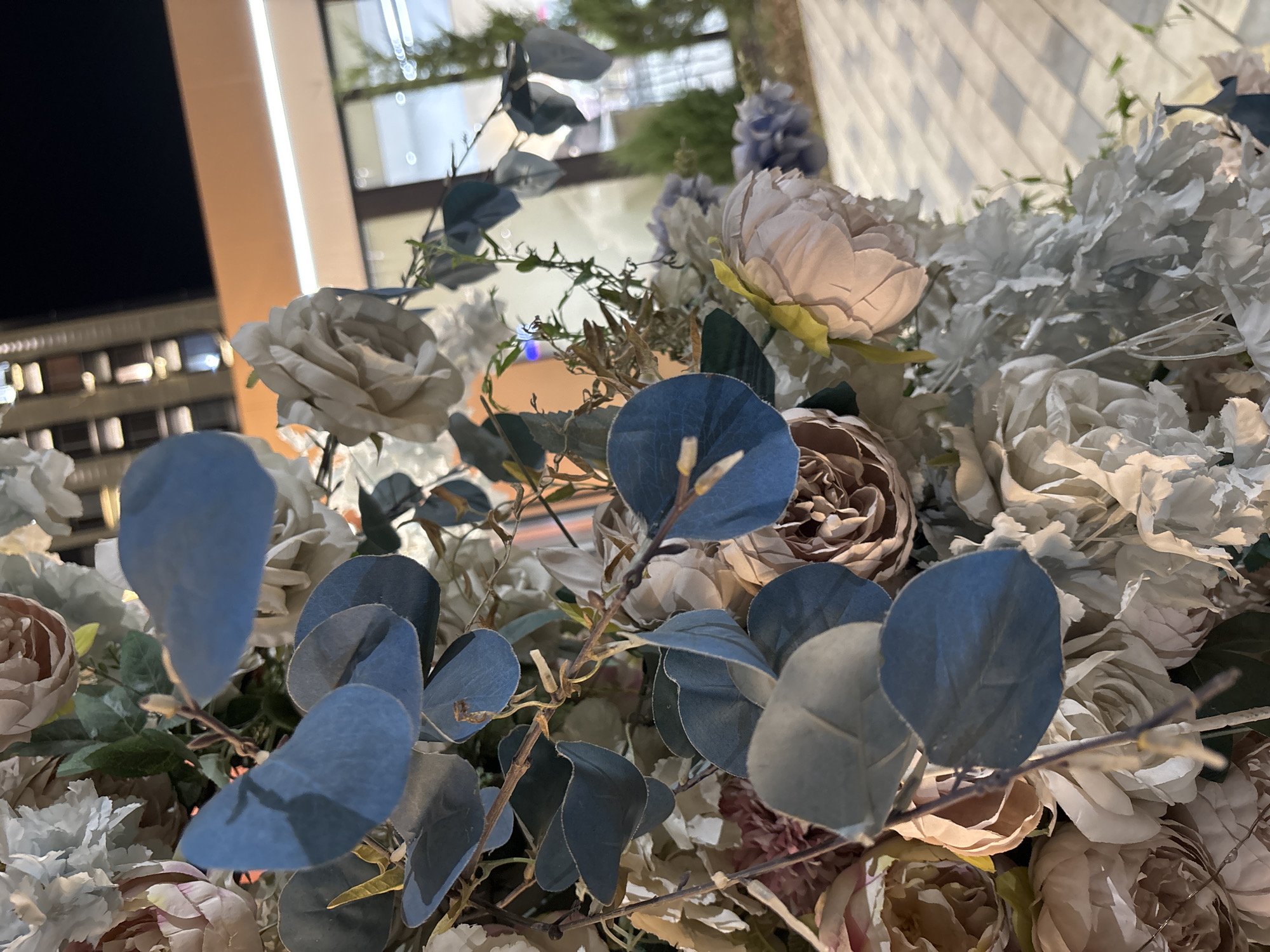 The image captures a close-up view of a floral arrangement, featuring a mix of pale pink, cream, and light blue artificial flowers, interspersed with blue-green eucalyptus leaves. The arrangement is positioned outdoors at night, with a modern building facade and illuminated windows visible in the blurred background. The building's architecture suggests an urban environment, possibly a commercial or residential complex. The ground beneath the arrangement appears to be paved with light-colored tiles, creating a patterned surface. The overall impression is one of a decorative element in an urban setting, likely part of an outdoor event or establishment. The image does not contain any people or animals. The exact location within the city of Shymkent, Kazakhstan cannot be determined from the visual information alone.