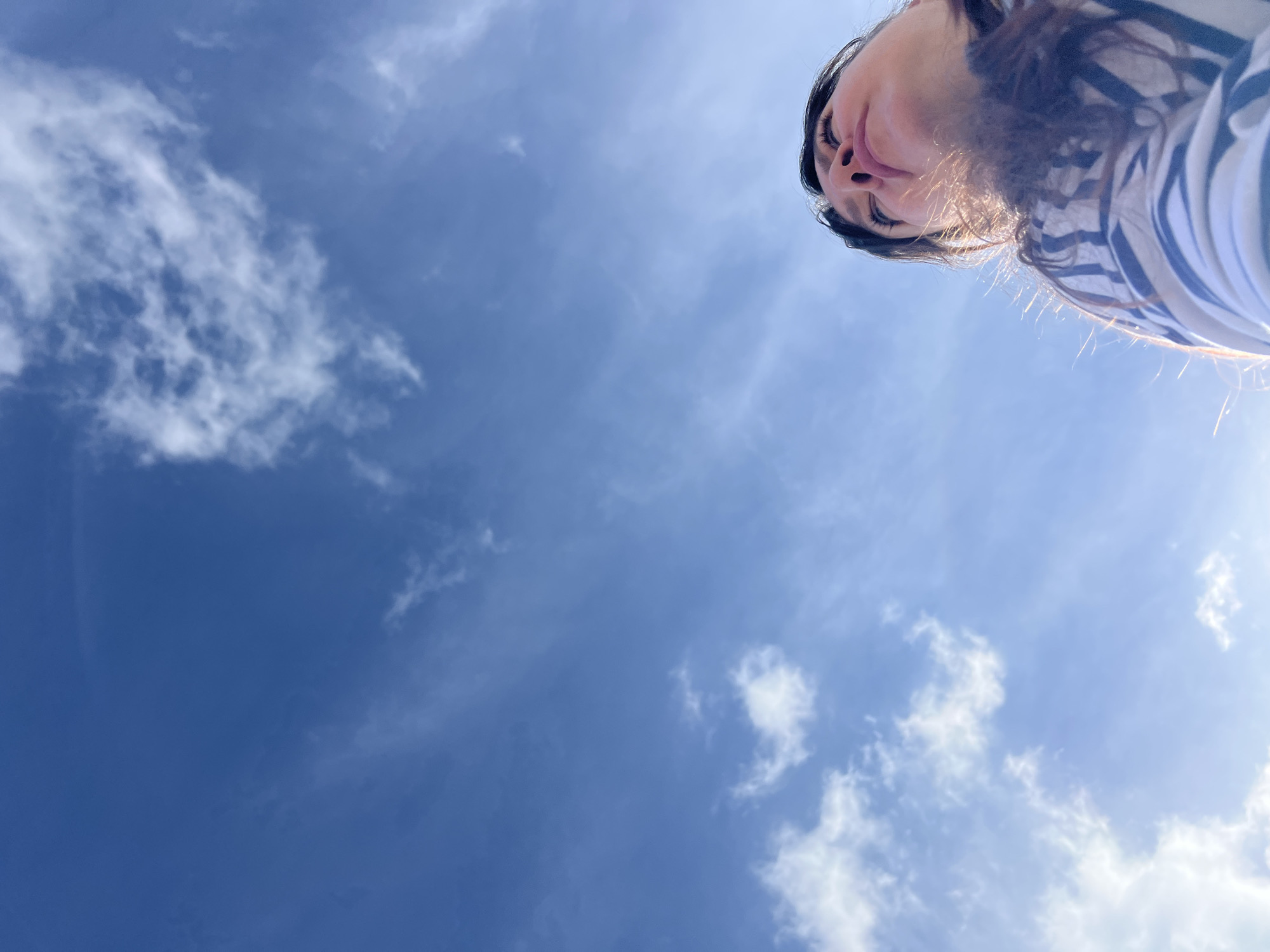 The image shows a low-angle shot of a woman looking up at a bright blue sky with wispy clouds. She is wearing a striped shirt. The scene appears to be outdoors, possibly in Noviy, Russia, though this is not explicitly visible in the image. The focus is on the woman's face and the expansive sky, with the bottom right corner of the image cropped to show her head and shoulders.