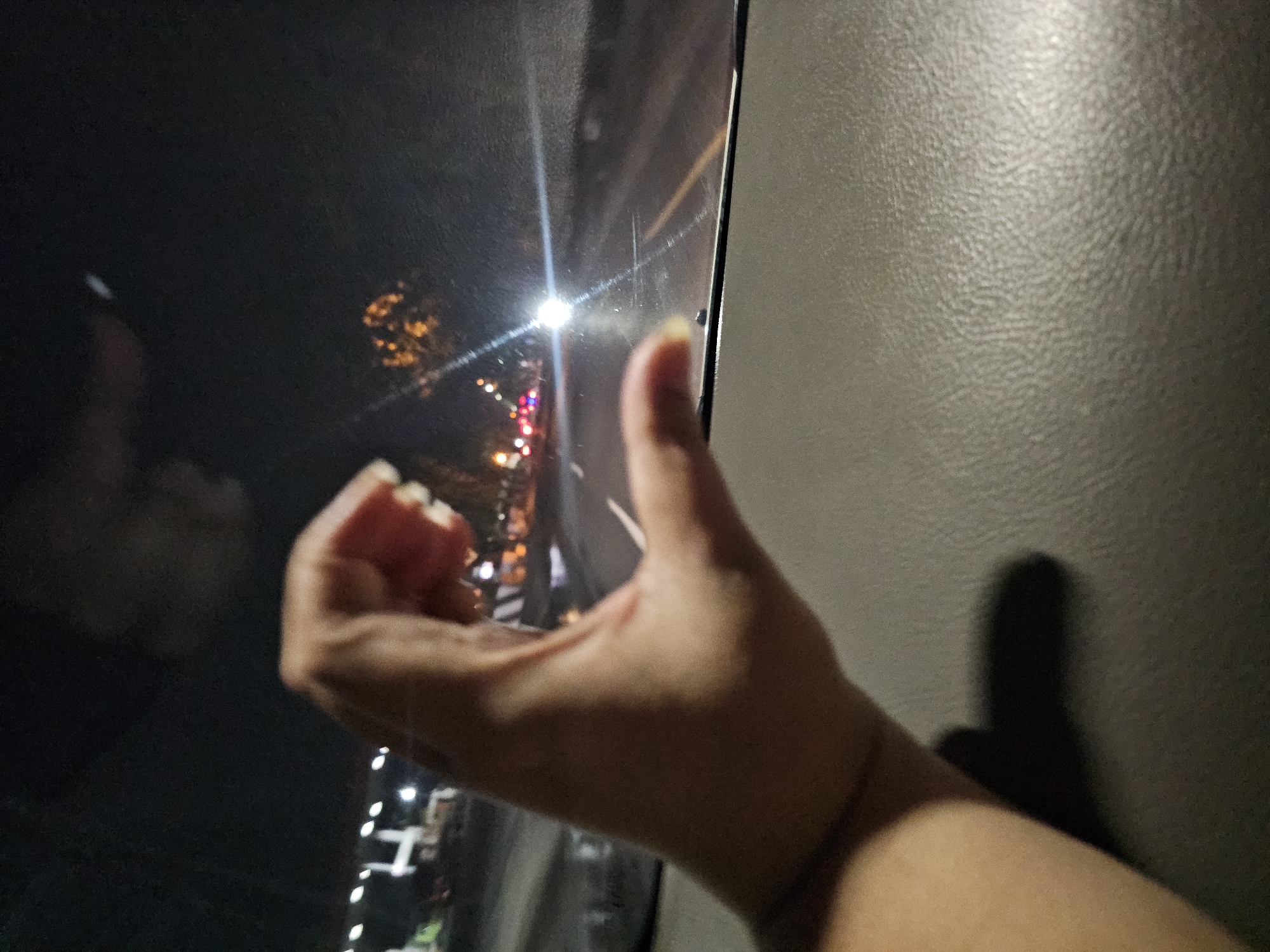 A hand is making a heart shape against the backdrop of a nighttime street scene in Palang, Indonesia. The hand, with fingernails painted, is in the foreground, its thumb and index finger creating the curves of a heart. In the mid-ground, through a car window, a street is visible with illuminated buildings and streetlights. A bright light, likely from oncoming traffic, creates a lens flare effect. The bottom portion of the image shows the textured surface of a car's dashboard. The overall impression is a candid, personal moment captured during a drive at night.