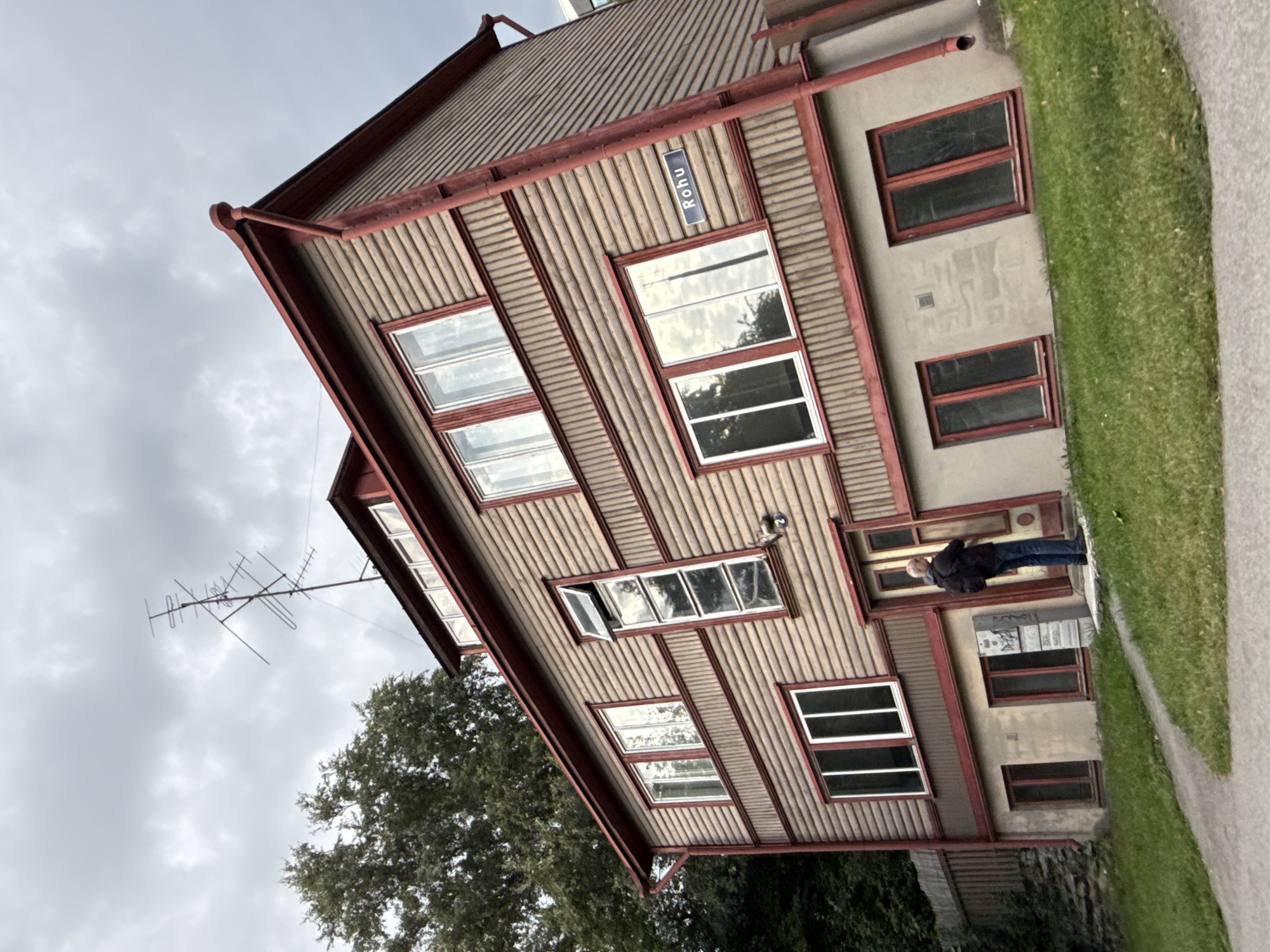The image captures a tilted perspective of a wooden house with red trim, situated on a grassy slope in Tallinn, Estonia. A dark-haired man wearing a black jacket and blue jeans stands at the entrance of the house, with his back to the camera, facing the door. The house has multiple windows, some of which reflect the cloudy sky. A blue street sign reading "Rõhu" is affixed to the wooden facade. A large antenna is mounted on the roof of the house, and dense foliage from a tree is visible to the left. The ground in front of the house is a mixture of paved walkway and green grass. The sky is overcast with grey clouds.