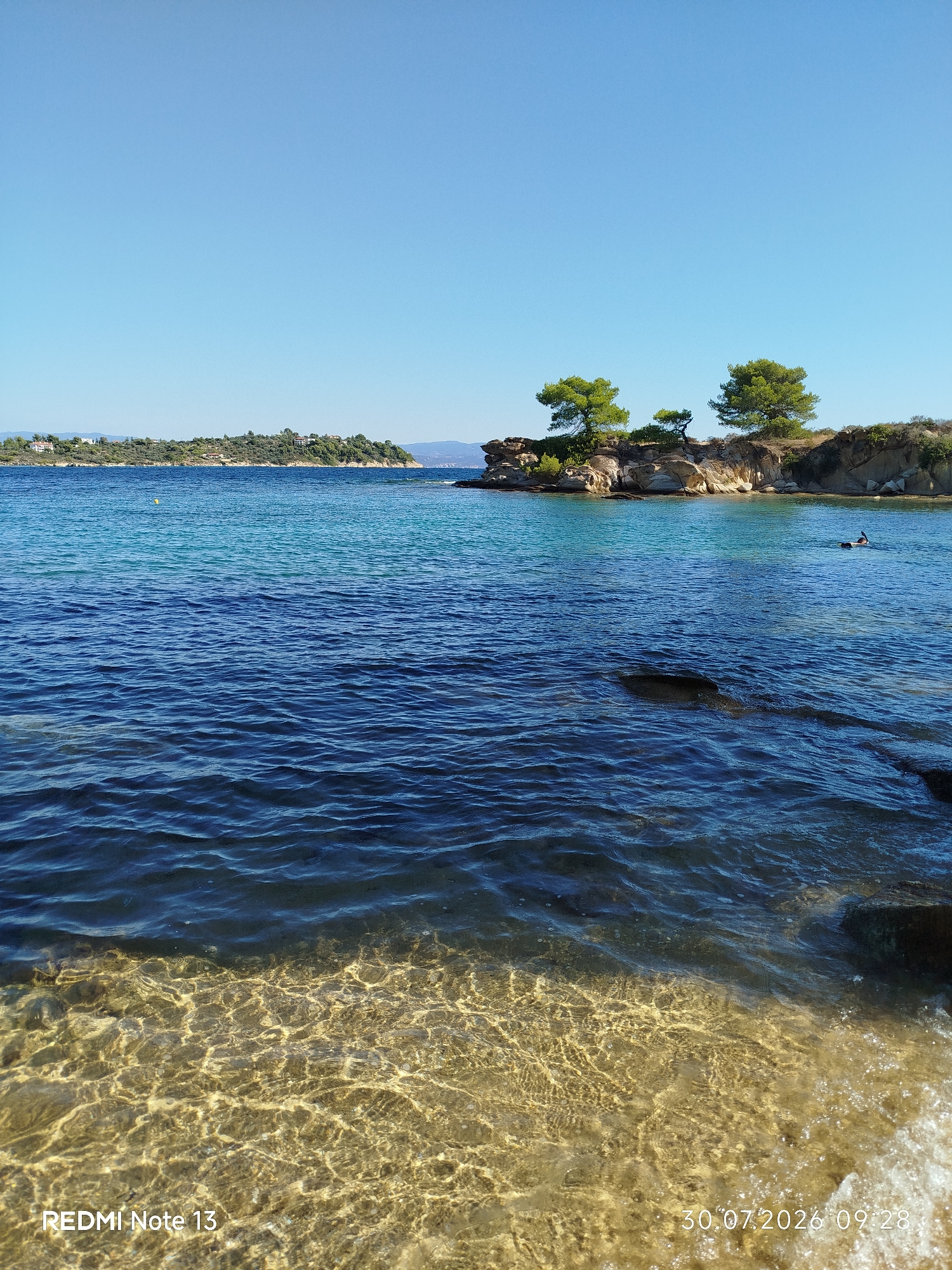 This is a vertical, eye-level, medium shot captured on a sunny day in Sithonia, Greece, featuring a serene coastal landscape. In the foreground, crystal-clear turquoise water laps onto a sandy beach, revealing the seabed with sunlight creating a shimmering effect. Further out, the deep blue ocean stretches towards a rocky outcrop dotted with evergreen trees. On the right side of this outcrop, a solitary individual is engaged in water activity, appearing to be snorkeling or swimming with fins. In the distance, a lush green peninsula with scattered buildings is visible under a clear blue sky. The bottom left of the image displays the text "REDMI Note 13" and the bottom right corner shows "30.07.2026 09:28".