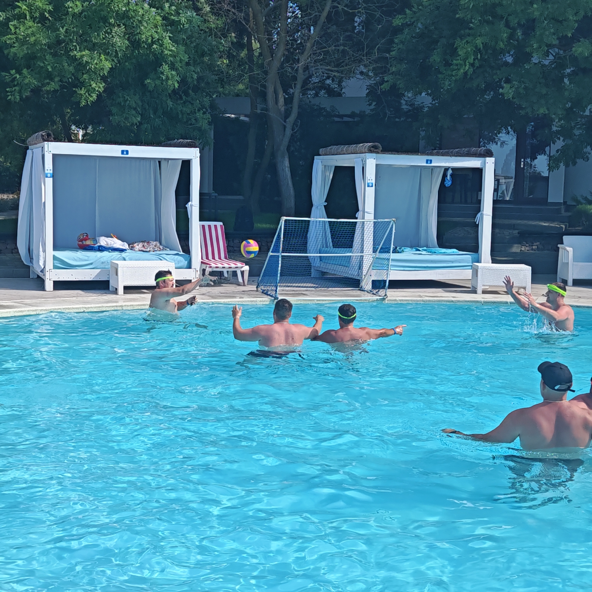 In a bright, sunlit outdoor pool, a group of men are engaged in a game of water polo. The clear blue water reflects the sunlight, creating a vibrant atmosphere. In the background, white cabana-style structures with draped curtains line the edge of the pool, suggesting a resort or hotel setting. A soccer-style goal net is set up in the water, indicating the playful nature of the activity.
Several men are actively playing, with some in the foreground splashing and reaching for a colorful ball that hovers in the air. Two men in the middle of the frame are facing away from the camera, their bodies partially submerged in the water as they participate in the game. Another man, wearing a green swim cap, is visible in the mid-right of the frame, his arms raised as if to block a shot or make a save. Towards the bottom right, another man is partially visible from behind, wearing a black baseball cap.
The time of day appears to be daytime, judging by the strong sunlight and shadows. The weather seems pleasant and warm, suitable for swimming and outdoor recreation. While no specific text is clearly visible on the people or the immediate objects of play, small blue numbered tags are present on the cabana structures. The overall mood of the scene is energetic and fun, with the individuals appearing to be enjoying their leisure time together. The presence of lounge chairs and the cabanas suggest a relaxed vacation environment.