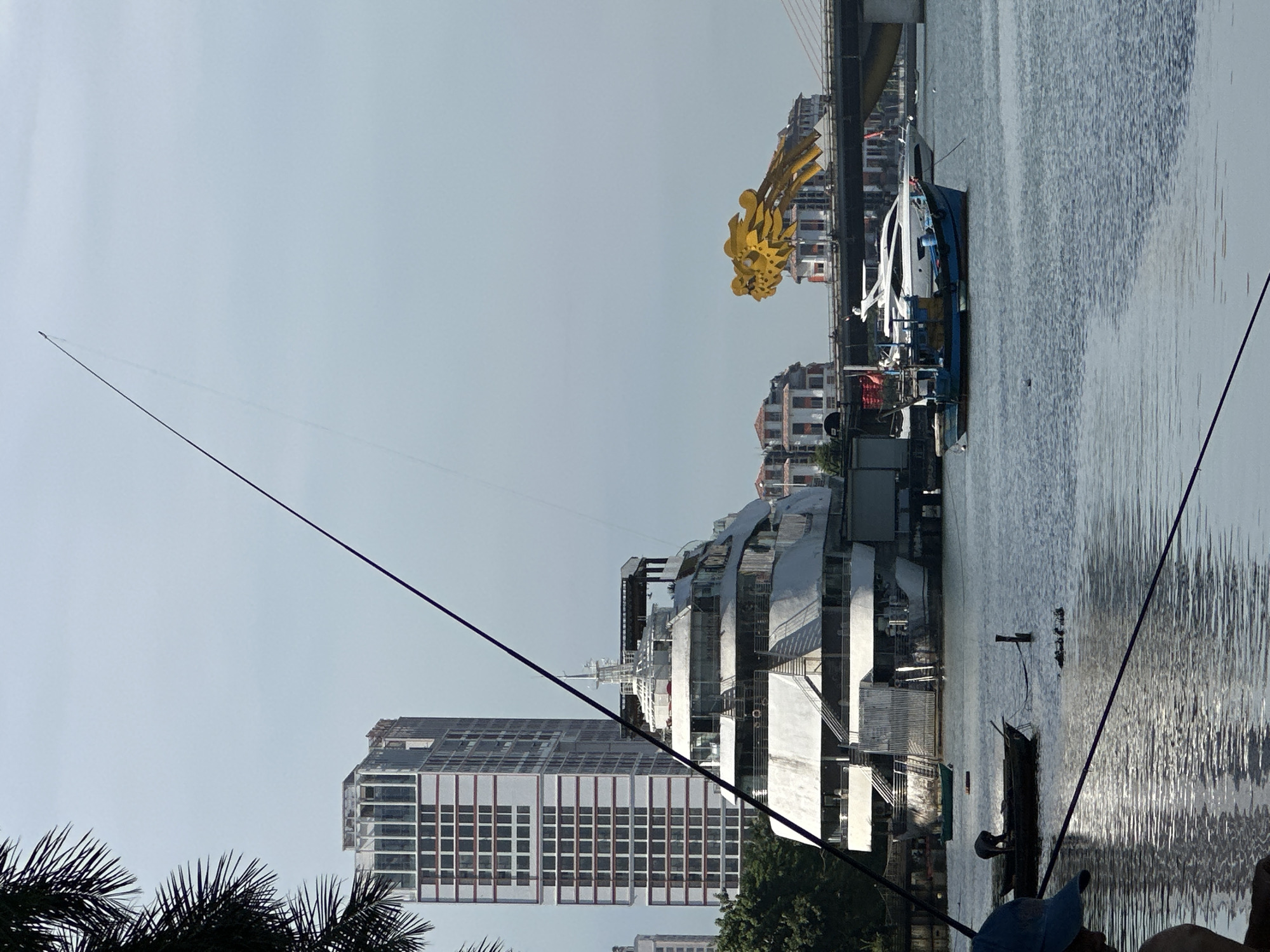 The image depicts a waterfront scene in Da Nang, Vietnam, likely along the Han River. Dominating the left side of the frame is a large, multi-story white vessel, possibly a tour boat or restaurant, with a distinctive tiered design. Behind it stands a tall, modern building with a red and white striped facade, characteristic of urban development in Da Nang. In the background, the iconic yellow dragon-shaped sculpture of the Dragon Bridge spans the river, with a section of its steel structure visible.
Several boats are present on the water. A small, dark fishing boat with a person tending to it is in the foreground. Further back, a blue and white motorboat is docked next to a larger, sleek white yacht. Two fishing rods, one extending high into the sky and another diagonally across the lower part of the frame, indicate recreational fishing activity. The water is calm, reflecting the sky and the surrounding structures. The sky is overcast, with a hazy light. Trees line the riverbank in the mid-ground. The overall impression is a blend of urban architecture, marine activity, and natural elements.