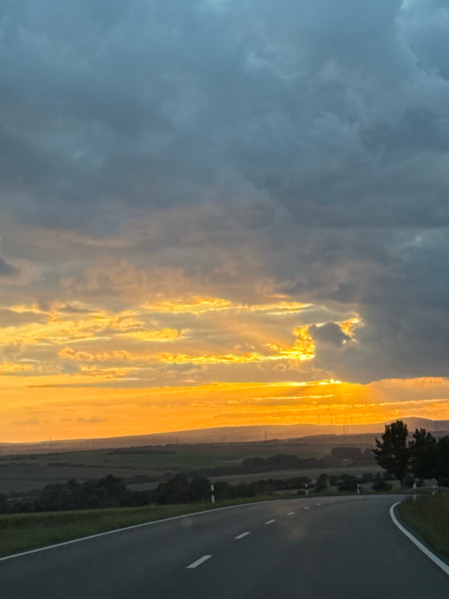 A paved road curves through a rural landscape under a dramatic sky. The road, with white lane markings, dominates the foreground and leads the viewer's eye towards the horizon. The sky is a striking contrast of dark, moody clouds in the upper portion and vibrant bands of yellow and orange sunlight breaking through lower down, suggesting either sunrise or sunset. The landscape beyond the road consists of rolling hills covered in fields and trees. In the middle distance, there are hints of human infrastructure, possibly power lines or distant buildings, interspersed with the natural environment. Two trees are visible on the right side of the frame, silhouetted against the bright sky. The scene is devoid of any people or animals, presenting a tranquil yet dynamic natural vista. The specific location is identified as Greußen, Germany.