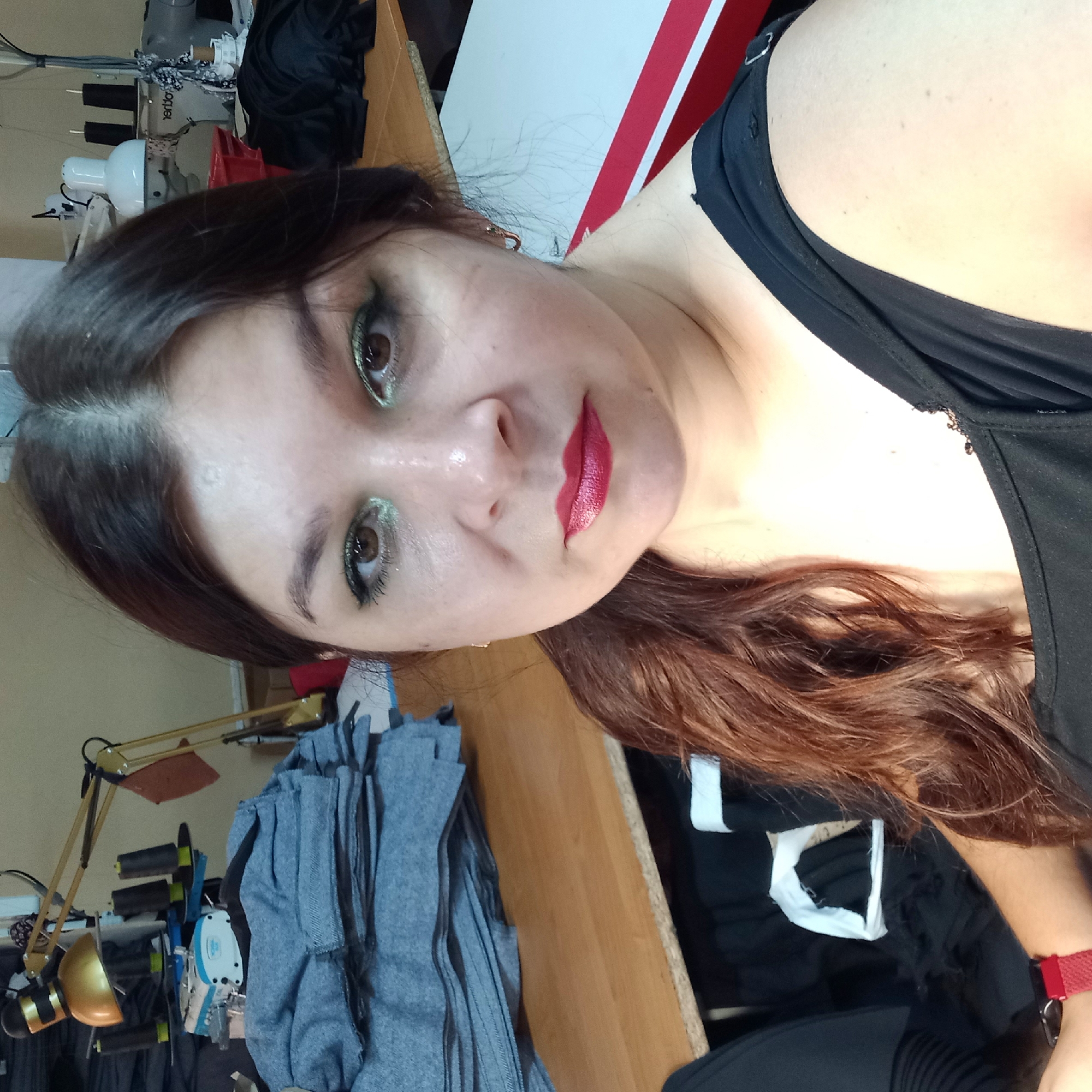 The image shows a medium shot of a woman in a sewing workshop. She has dark hair pulled to the side and is wearing a black top. Her makeup includes green eyeshadow and red lipstick. The background features sewing machines, stacks of fabric, and a desk with spools of thread and a lamp. The scene is likely indoors, possibly a home or small business, given the personal nature of the selfie and the cluttered but functional workspace. No specific details about Kropyvnytskyi, Ukraine are discernible from the image itself.
