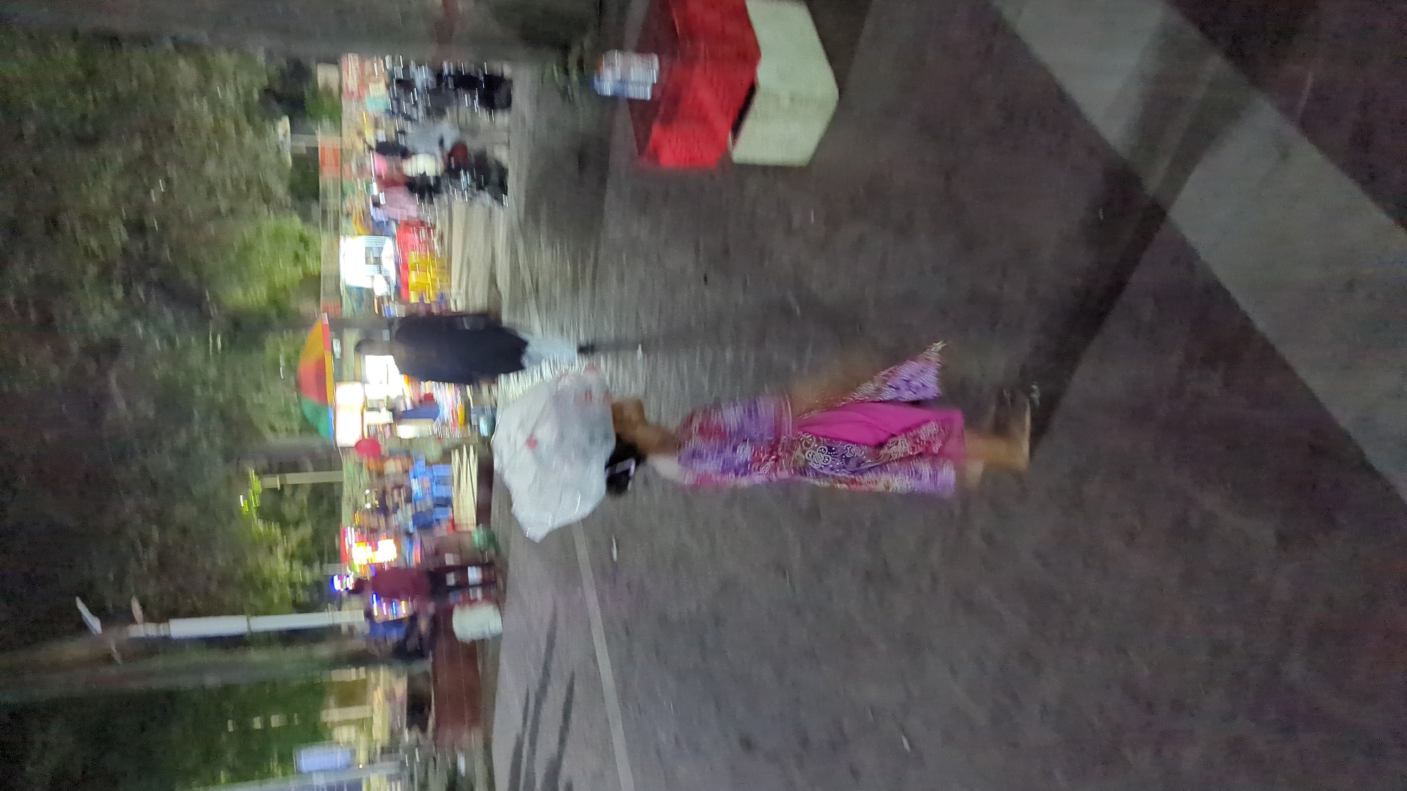 A child in a pink and purple patterned garment carries a large white bag of goods balanced on their head while walking on a paved pathway at night. The pathway is illuminated by artificial lights, and trees line the background, suggesting an outdoor urban setting. Several other individuals and food stalls are visible in the midground, indicating a busy public area. The scene is likely in Dhaka, Bangladesh, given the cultural attire and general ambiance often found in such locations.