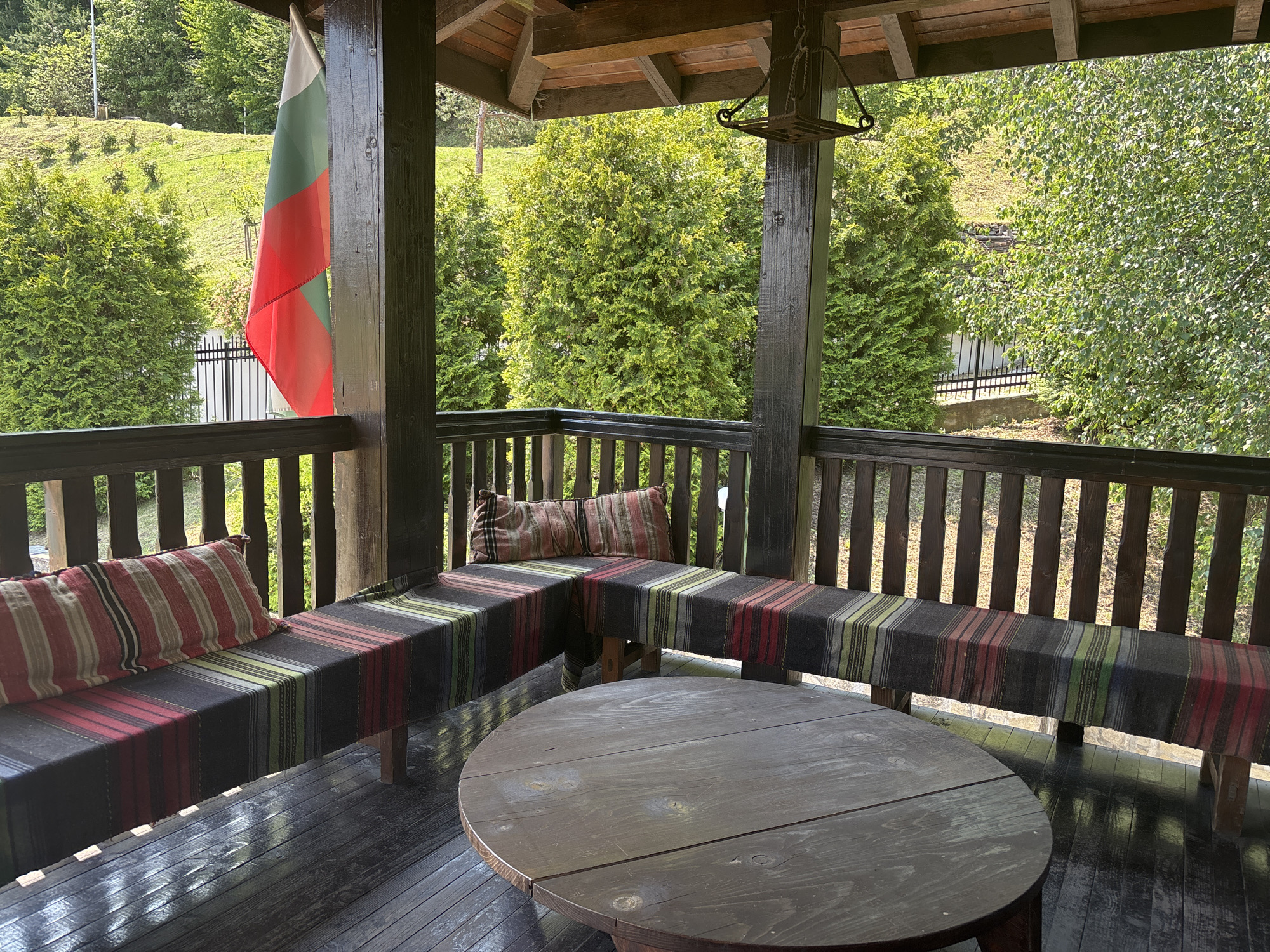 This image depicts a covered outdoor wooden porch or balcony situated in Belchin, Bulgaria. No individuals are present within the frame. The structure consists of dark-stained wooden posts, railings, and horizontal floorboards that exhibit a reflective sheen. A Bulgarian flag, displaying vertical white, green, and red stripes, is suspended from a wooden post on the left side of the frame. The porch ceiling features exposed wooden beams and planks, from which a dark, rustic wooden fixture, resembling a lantern, hangs.
An L-shaped wooden bench is built into the corner against the railing. It is covered with a dark textile adorned with multiple vertical stripes in colors including red, green, light brown, and grey. Two throw pillows, featuring a striped pattern predominantly in red, white, and black, are placed on the bench seats. A round, dark-stained wooden table occupies the central foreground of the porch.
Beyond the porch railing, the background reveals a natural landscape dominated by lush green vegetation. This includes dense evergreen trees, a deciduous tree with vibrant green foliage, and a grassy, upward-sloping hillside. Sections of a black metal fence are visible beneath the trees and along the lower edge of the vegetated area.