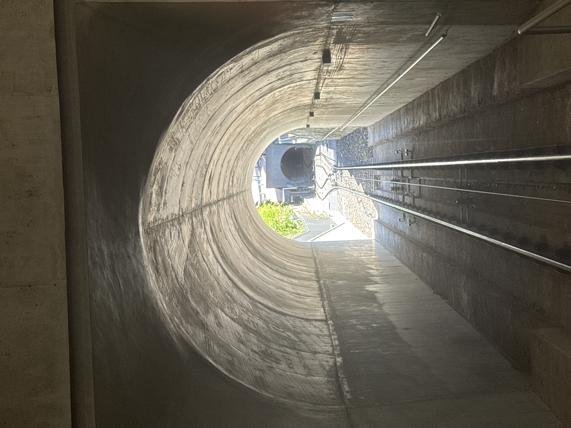 This is a low-angle, eye-level shot from inside a concrete tunnel looking out towards a second, smaller tunnel entrance. The tunnel has a curved, arched ceiling and rough concrete walls. Two parallel sets of train tracks run down the center of the tunnel, converging towards the exit. On either side of the tracks, there are concrete platforms and inclines. There are metal handrails along the right-hand wall. Outside the tunnel, sunlight illuminates lush green foliage and the entrance to the next tunnel. The scene appears to be the funicular tunnel leading to the station at Monte San Salvatore in Lugano, Switzerland. There are no people visible in the image.