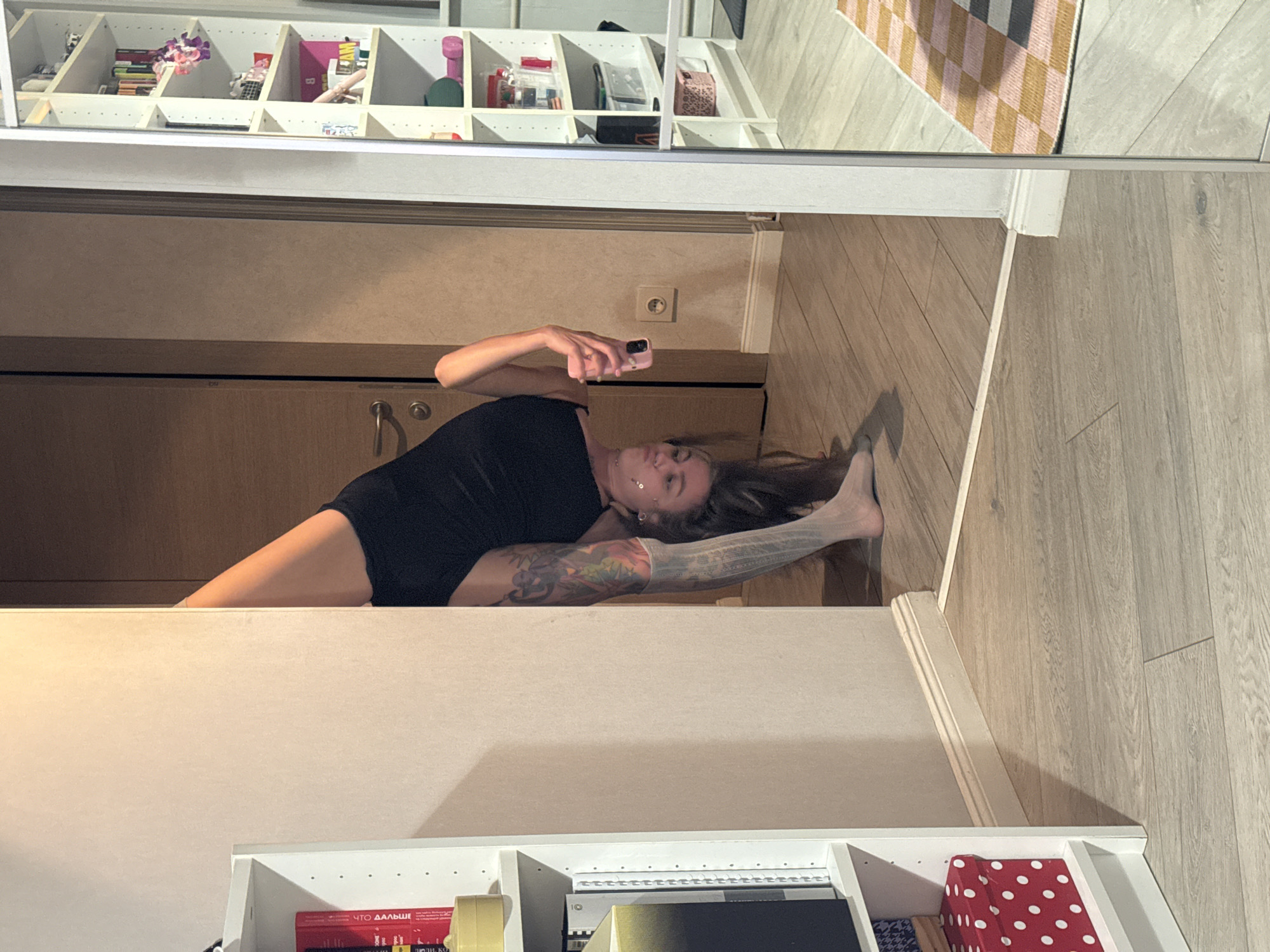 A woman is in a handstand in a dimly lit room, possibly a living room or bedroom, indoors. She is wearing black shorts and a black top, with white socks. She has tattoos on her legs and arms. She is holding a pink phone in her right hand. In the background, there is a doorway, a tall white bookshelf filled with books and items, and a mirror that reflects a portion of the room, including a rug with a checkered pattern. The floor is made of light-colored wood. The scene suggests a domestic environment. There is no indication of the city of Moscow, Russia in the image.
