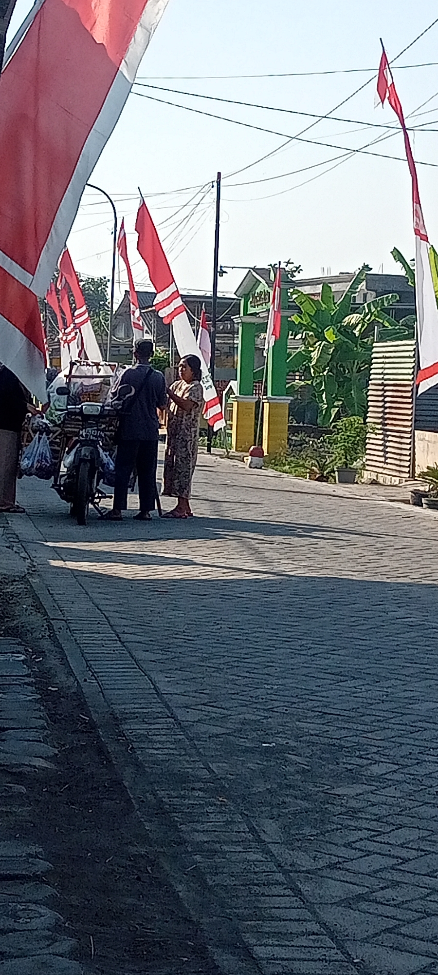 The image depicts a street scene in Gempolklutuk, Indonesia, under a clear sky. Several red and white flags, some with a white stripe at the bottom, are prominently displayed, suggesting a celebration or national event. In the foreground, a person is partially visible on the left, wearing dark clothing. To the right of them, a motorcycle is parked, loaded with items in plastic bags. Two individuals are standing near the motorcycle, engaged in conversation. One is a man wearing a dark t-shirt and a cap, facing the motorcycle. The other is a woman wearing a patterned dress, facing the man. In the background, a green arched gateway with yellow pillars is visible, adorned with flags. Beyond the gateway, buildings and lush green vegetation, including banana trees, are present. The street is paved with interlocking concrete blocks, and shadows are cast across the surface, indicating a bright, sunny day. The overall atmosphere appears to be one of everyday life augmented by festive decorations.