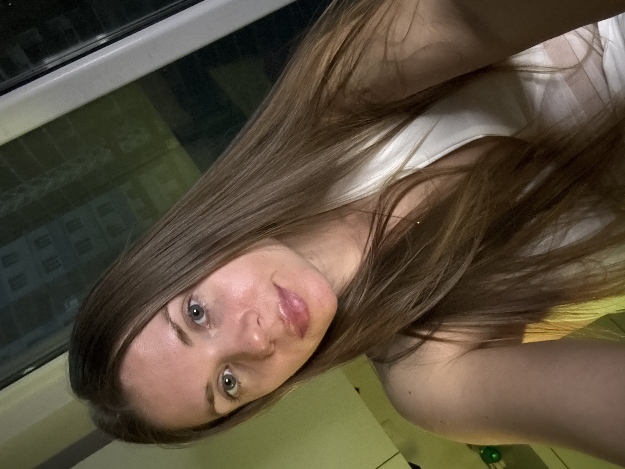 A selfie captures a young woman with long, straight, light brown hair flowing over her shoulders, her head tilted. She looks directly at the camera with clear blue-gray eyes and a soft, neutral expression, her lips slightly glossy. She is wearing a light-colored, possibly white, sleeveless top. The setting appears to be indoors, likely in Batumi, Georgia, beside a window. Through the window, or reflected in it, faint outlines of urban buildings are visible against a dim background, suggesting it is evening or night. The overall lighting is soft and ambient. No text is visible in the image.