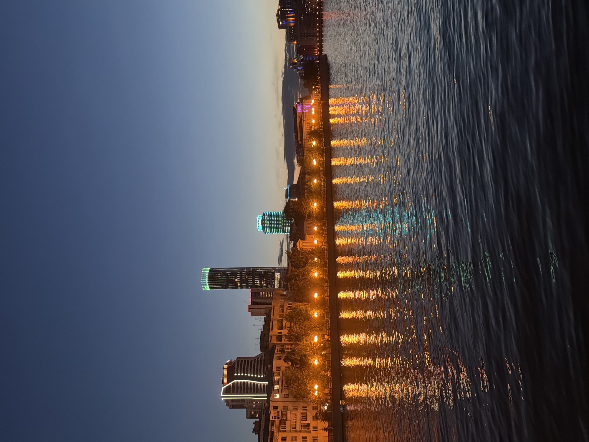 This is a nighttime view of the Yekaterinburg city skyline, as seen from across the Iset River. The image captures a cluster of illuminated buildings, including several modern skyscrapers, contrasted against a darkening sky. The most prominent skyscraper is a tall, cylindrical building with green lights around its top and a pattern of white lights down its facade. To its left is another skyscraper with a distinctive geometric design outlined in white lights. In the foreground, the surface of the Iset River reflects the lights from the city, creating streaks of orange and blue light on the dark water. Along the riverbank, a row of trees and lampposts are visible, also illuminated and casting reflections. The overall impression is of a vibrant urban landscape at dusk or early evening, with the city's lights beginning to dominate the scene.