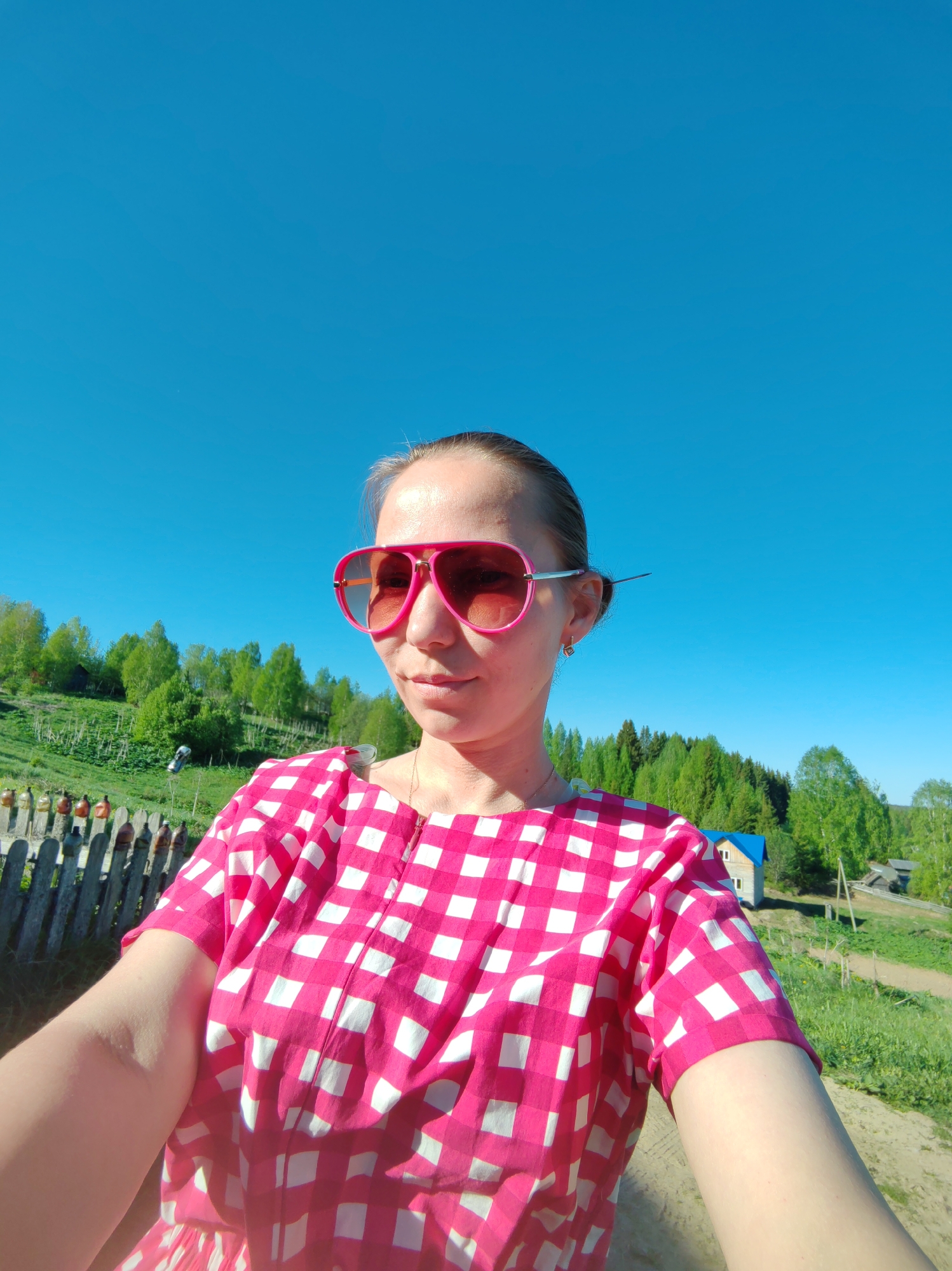 A woman wearing pink sunglasses and a pink and white checkered dress is taking a selfie. The background features a bright blue sky, a green hillside with trees, a wooden fence, and a small house with a blue roof. The woman is in Чухлэм, Russia.