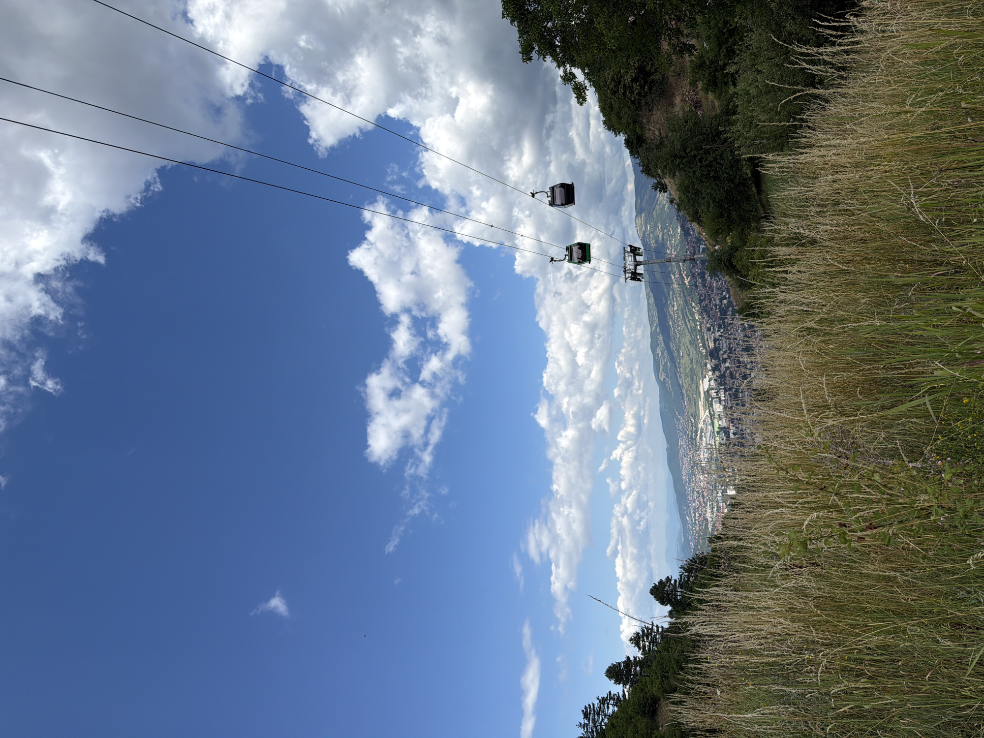 This is a vertical, low-angle shot of cable cars moving against a blue sky dotted with white clouds. The cable cars are suspended from thin wires that stretch across the frame from left to right. There are at least three visible cable cars, two with dark colored cabins and one with a green cabin.
Below the cable cars, a city is visible in the distance, nestled in a valley. The city appears to be comprised of many buildings with red roofs. The foreground is dominated by tall, dry grass, giving a sense of being on a hillside. A dark green forest is also visible on the right side of the image, bordering the grassy slope.
The scene evokes a sense of travel and a panoramic view. The bright sky and scattered clouds suggest fair weather, likely daytime. The presence of the city and the cable car system indicates an urban or semi-urban setting, possibly a tourist attraction or a mode of transportation in a hilly or mountainous area, consistent with Sarajevo. No text is visible in the image.