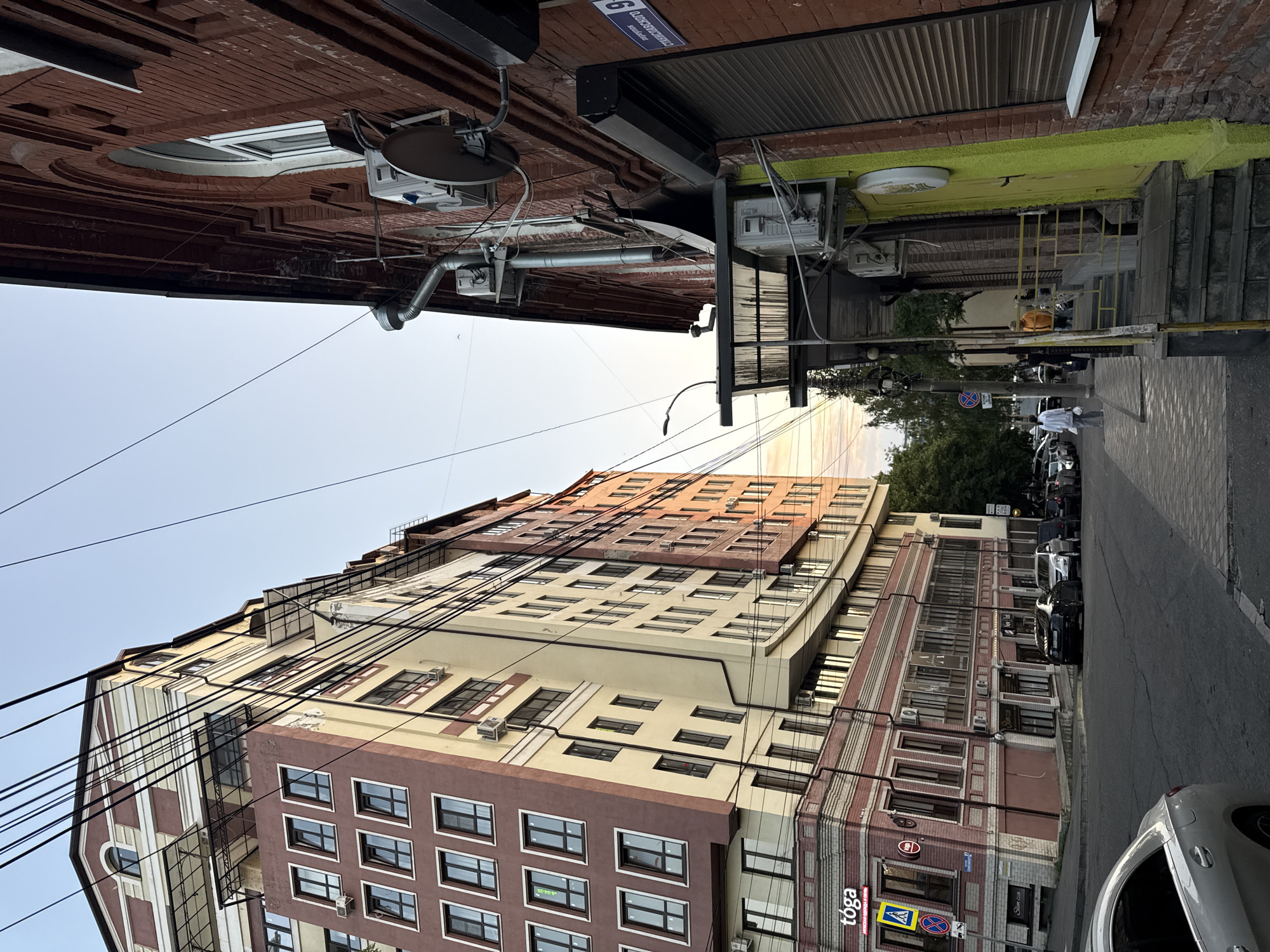 This is a low-angle, vertical shot of a city street in Sulak, Russia. The scene depicts a mix of old and new architecture, with several multi-story buildings lining the street. The sky is clear and appears to be either early morning or late afternoon, judging by the soft lighting.
In the foreground, a silver car is parked on the right side of the street, its rear end visible. The street itself is paved, with a sidewalk on the right. Along the sidewalk, there are several parked cars, including a black SUV. A few people are visible walking or standing near the buildings, dressed in casual clothing. One person in a white shirt and grey pants is walking away from the camera.
The buildings are varied in their design. On the left, a taller, modern building features a light-colored facade with many windows and visible air conditioning units. Further down the street, there are older brick buildings with decorative elements. Some of these buildings have storefronts at street level. Signs are visible on some of these storefronts, including one that reads "tōga" and another with a red circle and a blue cross inside a yellow diamond, indicating a traffic sign. Another sign appears to say "Slim Club."
The upper parts of some buildings show exposed brickwork and architectural details. Electrical wires are strung across the frame, adding a sense of urban density. The overall impression is a typical urban street scene in Russia, showcasing a blend of historical and contemporary architecture under a clear sky.