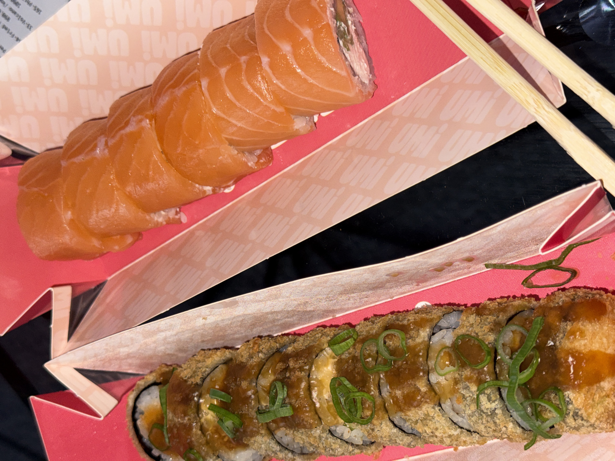 The image shows a take-out sushi order, likely from the restaurant Umi in Belgrade, Serbia, given the branding on the container. A row of salmon nigiri sushi is placed at the top, with overlapping slices of salmon covering the rice. Below, there is a row of tempura-fried sushi rolls drizzled with sauce and garnished with green onions. Chopsticks are visible next to the sushi. The sushi is served in a pink take-out container with the restaurant's name printed on it in a repetitive pattern. The background is black.