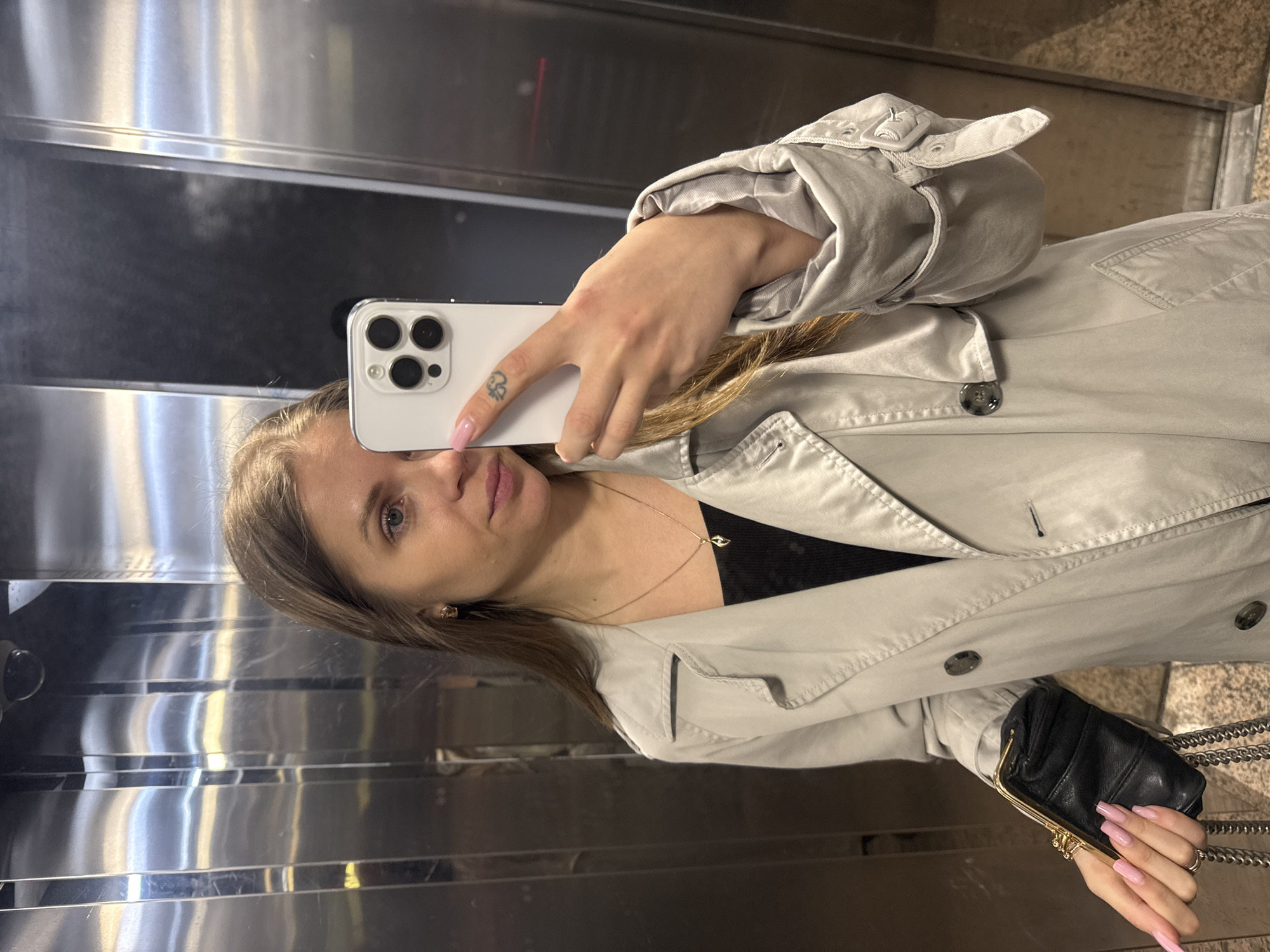 This vertical mirror selfie captures a woman inside what appears to be an elevator or a space with reflective, brushed metallic walls, typical of urban settings in Batumi, Georgia. The woman, with light brown hair and blue eyes, looks directly at the camera with a neutral expression.
She is wearing a light beige or grey trench coat over a black top, complemented by a delicate gold necklace with a small, dark pendant. She holds a white smartphone, identifiable by its triple camera array (likely an iPhone), in her left hand, partially obscuring her face. Her nails are long and painted a soft pink, and a small, abstract tattoo is visible on her left index finger. In her right hand, she carries a small black clutch purse with a gold-toned clasp and chain. A small gold stud earring is visible in her right ear.
The metallic walls of the setting reflect ambient light, creating varying shades and highlights across the surface. The image is well-lit, suggesting an indoor environment. No visible text is discernible in the image.