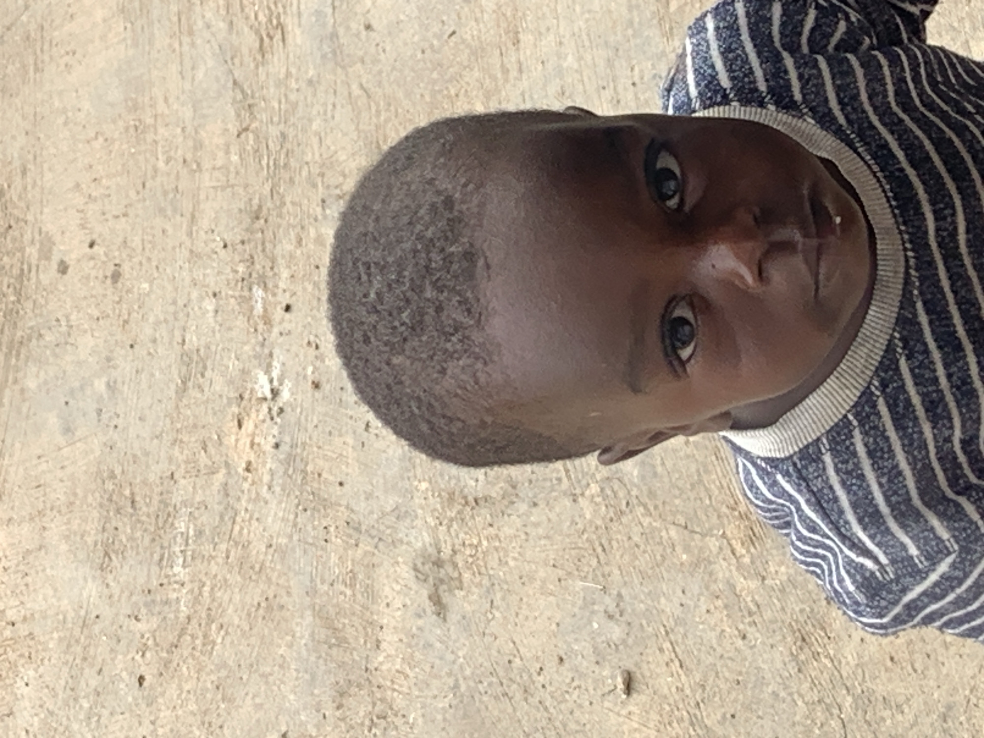 An upward shot frames a young Black child's face, wearing a blue and white striped shirt, against a neutral, textured background of what appears to be a dusty, concrete surface, common in urban areas such as Jos, Nigeria. The child's hair is short, and their gaze is directed at the camera. The lighting is bright, casting subtle shadows on the child's features, and a small white speck is visible on their lower lip. The background is out of focus, emphasizing the subject.