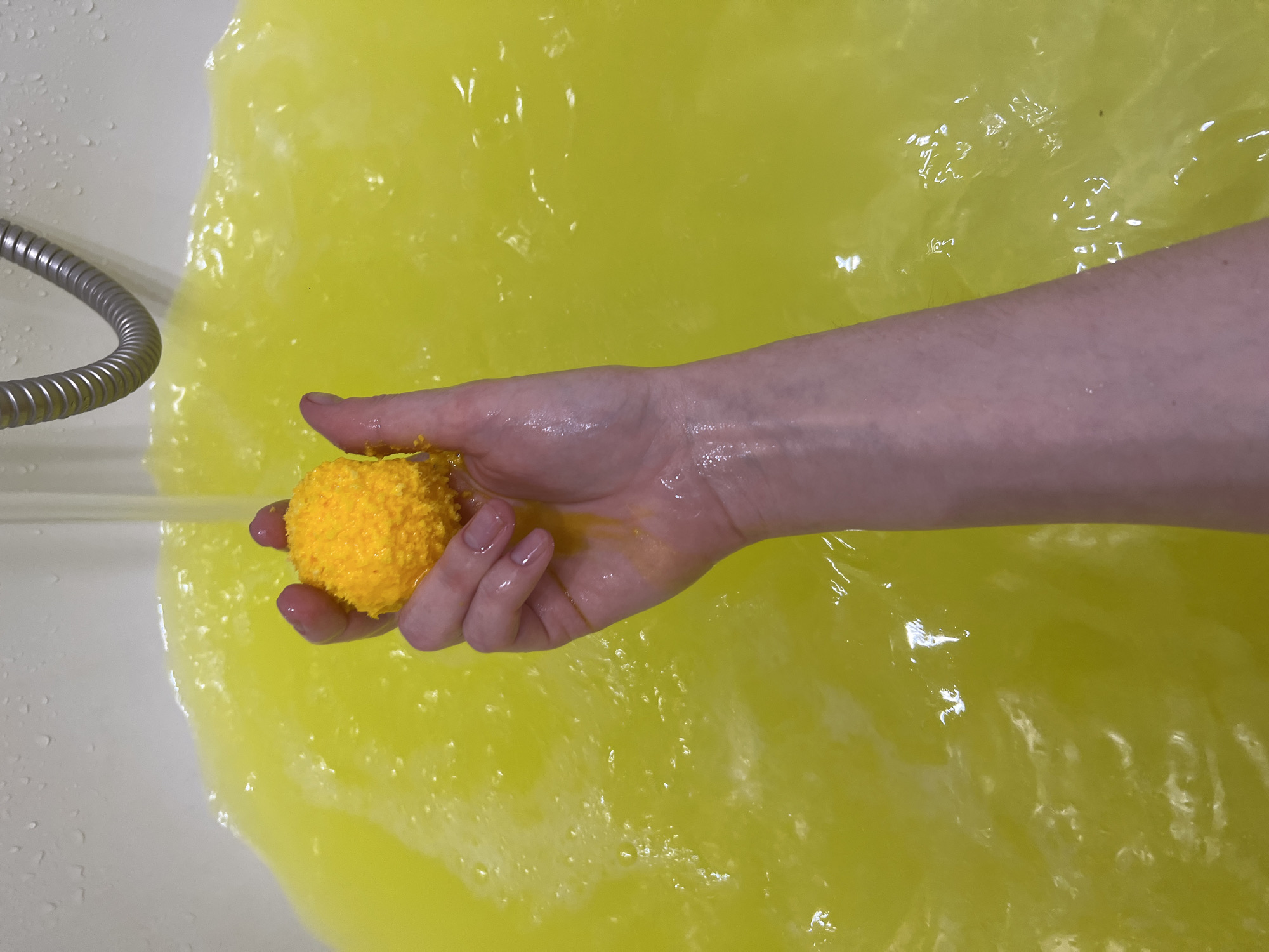 A high-angle shot depicts a person's hand holding a yellow bath bomb under running water from a showerhead in a bathtub filled with yellow water. The showerhead is chrome and flexible. The person's hand is pale. The location, Khimki, Russia, cannot be verified from the image.