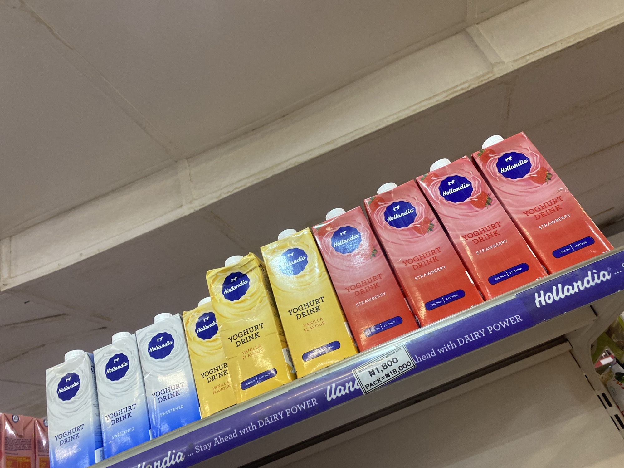 The image shows a shelf in a store, likely a supermarket or convenience store, displaying rows of Hollandia yoghurt drinks. The products are arranged in various flavors, including strawberry and vanilla. The shelf also has a price tag indicating "1,800 PACK=18,000" and a slogan that reads "...Stay Ahead with DAIRY POWER". The lighting suggests an indoor environment, possibly under fluorescent lights, and the shelves appear to be made of light-colored material. The background is out of focus but suggests a typical retail setting. The image was taken in Ikorodu, Nigeria.