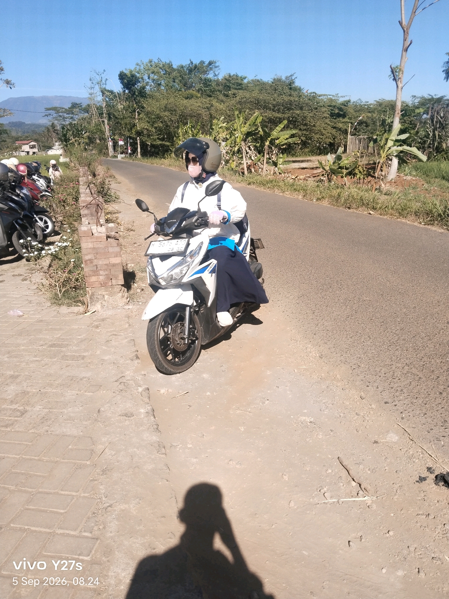 A woman wearing a helmet, sunglasses, a white jacket, and dark blue pants is seated on a white Honda scooter. The scooter has a license plate that reads "H 5676 ADO". She is on a paved road in what appears to be a rural area. To the left of the road, there is a dirt path and a row of parked motorcycles. Beyond the motorcycles, there is a brick structure and vegetation including trees and banana plants. In the background, there are more trees and a distant mountain. The sky is clear and blue. The timestamp "5 Sep 2026, 08.24" and "vivo Y27s" are visible in the lower left corner of the image. The location is identified as Selojetis, Indonesia.