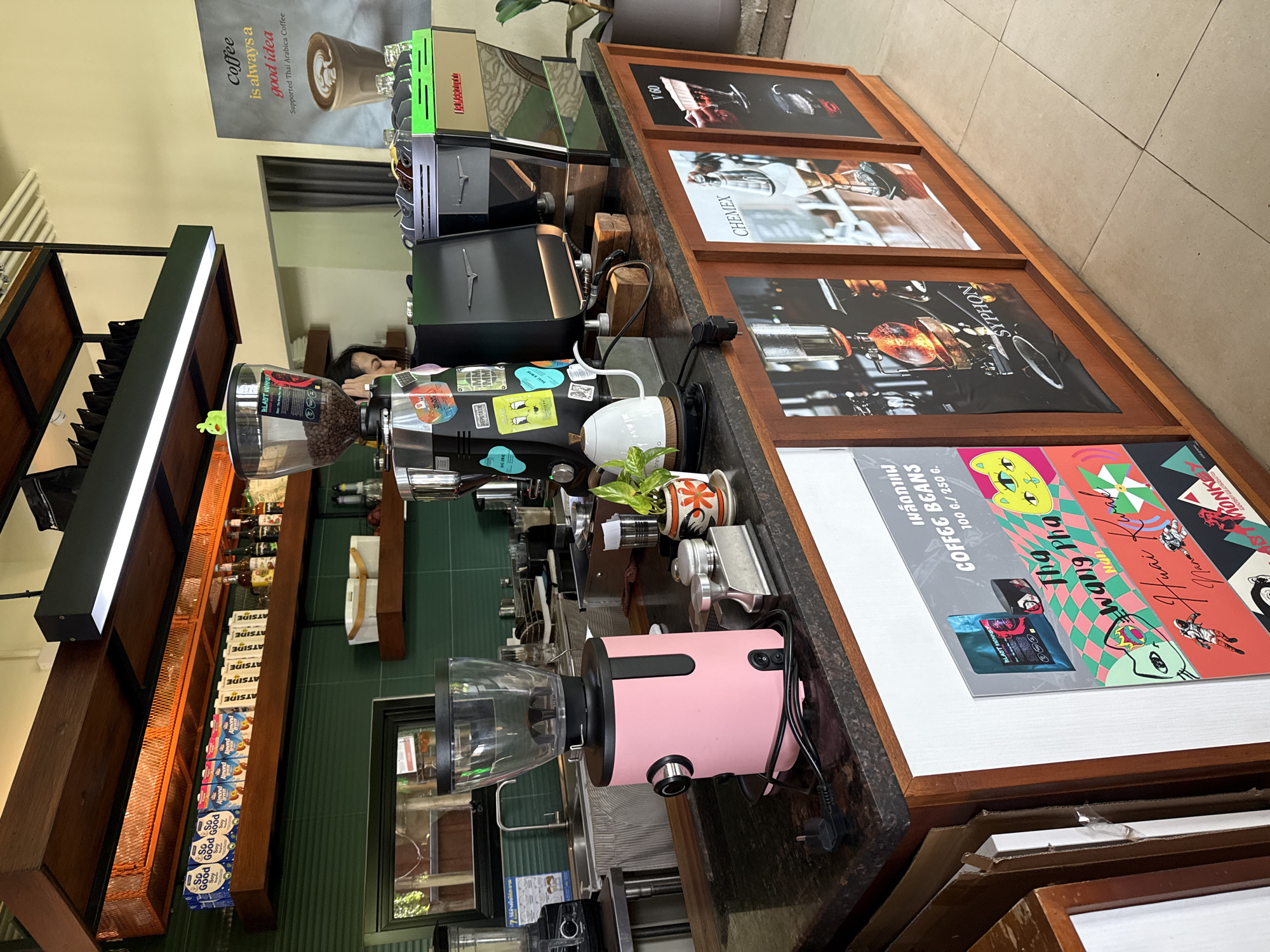 A brightly lit, modern coffee shop counter in Ko Samui District, Thailand, features a pink coffee grinder, a black espresso machine with a neon green trim, and a white electric kettle. Behind the counter, shelves are stocked with various coffee beans and beverages, including "So Good" soy milk cartons. A poster on the wall advertises "Coffee is always a good idea." The counter itself is decorated with framed artwork showcasing different coffee brewing methods like Chemex and Syphon. A barista is partially visible behind the espresso machine, attending to a customer. The shop appears clean and well-organized, with a focus on artisanal coffee.