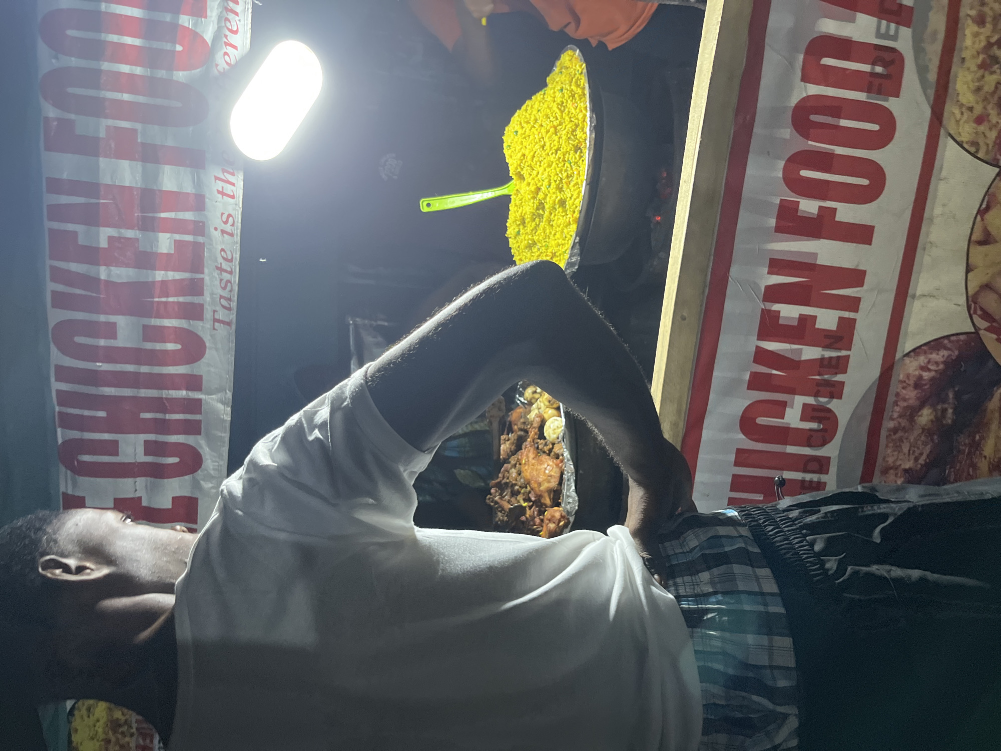 A young man in a white t-shirt and plaid shorts stands with his back to the camera, looking at food stalls. A banner above the stalls reads "CHICKEN FOOD". Below the banner, a large pot of bright yellow rice is being served with a green ladle. To the left, a platter of fried chicken and other food is visible. The scene is illuminated by a bright light, creating a dimly lit, outdoor atmosphere, likely a street food vendor.