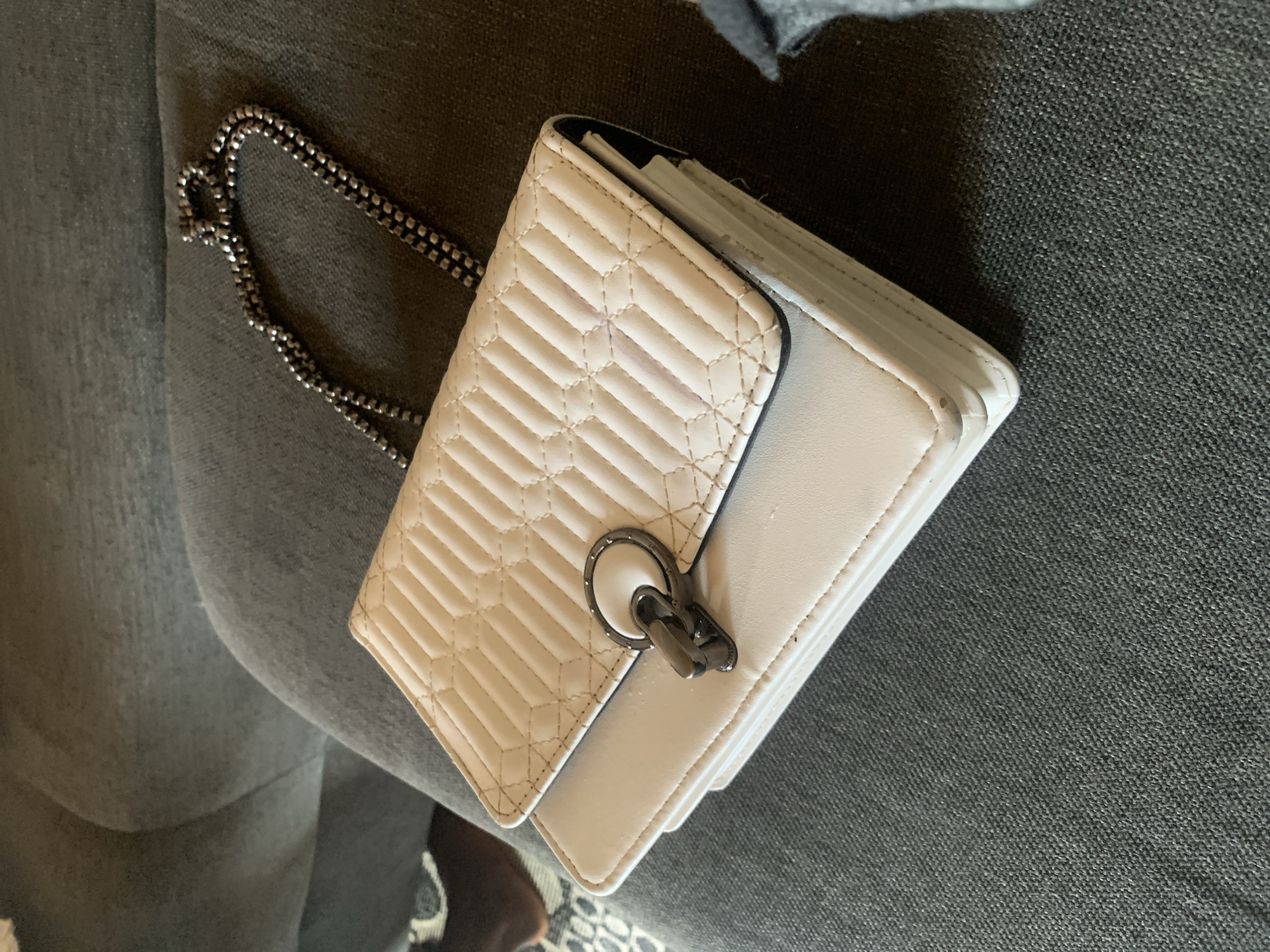 A light-colored, quilted handbag with a silver chain strap rests on a dark grey, textured couch. The handbag features a prominent metal clasp with a circular element and a rectangular latch. The couch appears to be in a residential setting. There are no visible people or indications of Sabon Tasha, Nigeria.