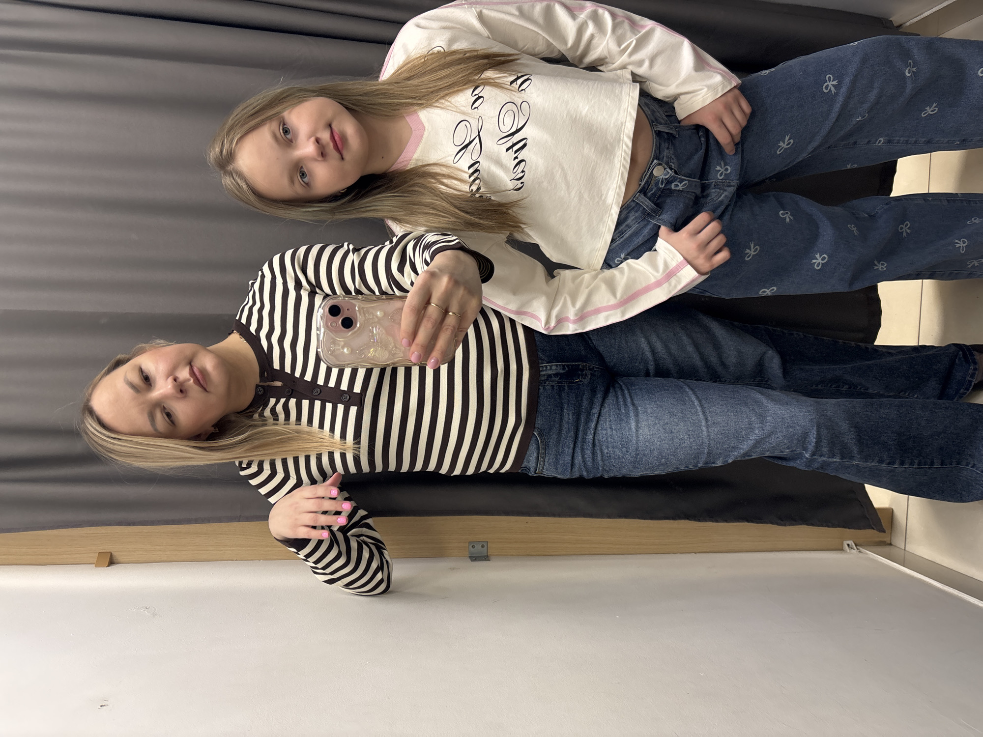 Two females are in a dressing room taking a selfie. The older female is wearing a black and white striped long-sleeved shirt and blue jeans. She is holding a phone in her right hand. The younger female is wearing a long-sleeved white shirt with pink trim and writing and blue jeans with bow patterns. They are sitting on a black bench in a room with a grey curtain and white walls. The location is not evident from the image.