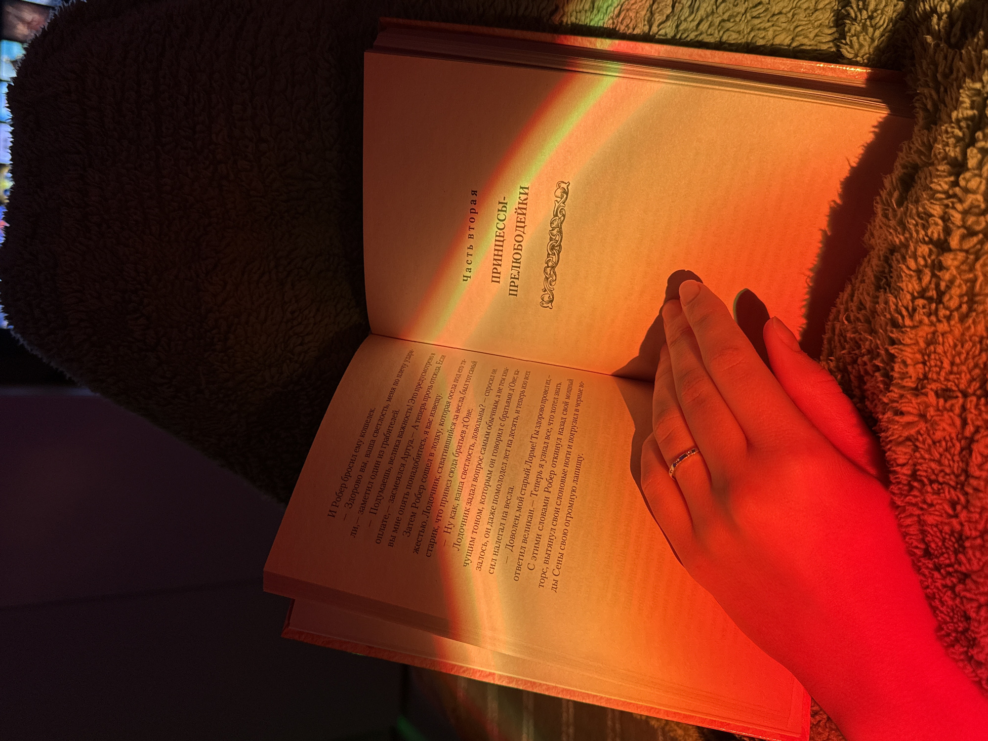 A close-up, high-angle shot captures an open book resting on a textured, dark green/brown blanket, illuminated by warm, colorful light. A hand, adorned with a ring on the ring finger, is gently placed on the right page of the book, suggesting someone is reading. A prominent rainbow-like light effect, featuring distinct bands of red, orange, yellow, and green, bathes the pages and the hand, creating a cozy and atmospheric glow. The lighting suggests either artificial light from a prism or lamp, or perhaps natural light filtered in a unique way. The background to the left is dark and indistinct, with subtle hints of colorful, blurred lights.
The visible text on the right page of the book is in Russian. The main title of the section reads "Часть вторая" (Part Two) and "ПРИНЦЕССЫ-ПРЕЛЮБОДЕЙКИ" (Princesses-Adulteresses). The body text describes a narrative involving characters like "Робер" (Robert) and a boatman, discussing money and past events. The overall impression is one of quiet contemplation or relaxed reading in a warmly lit, comfortable setting.
