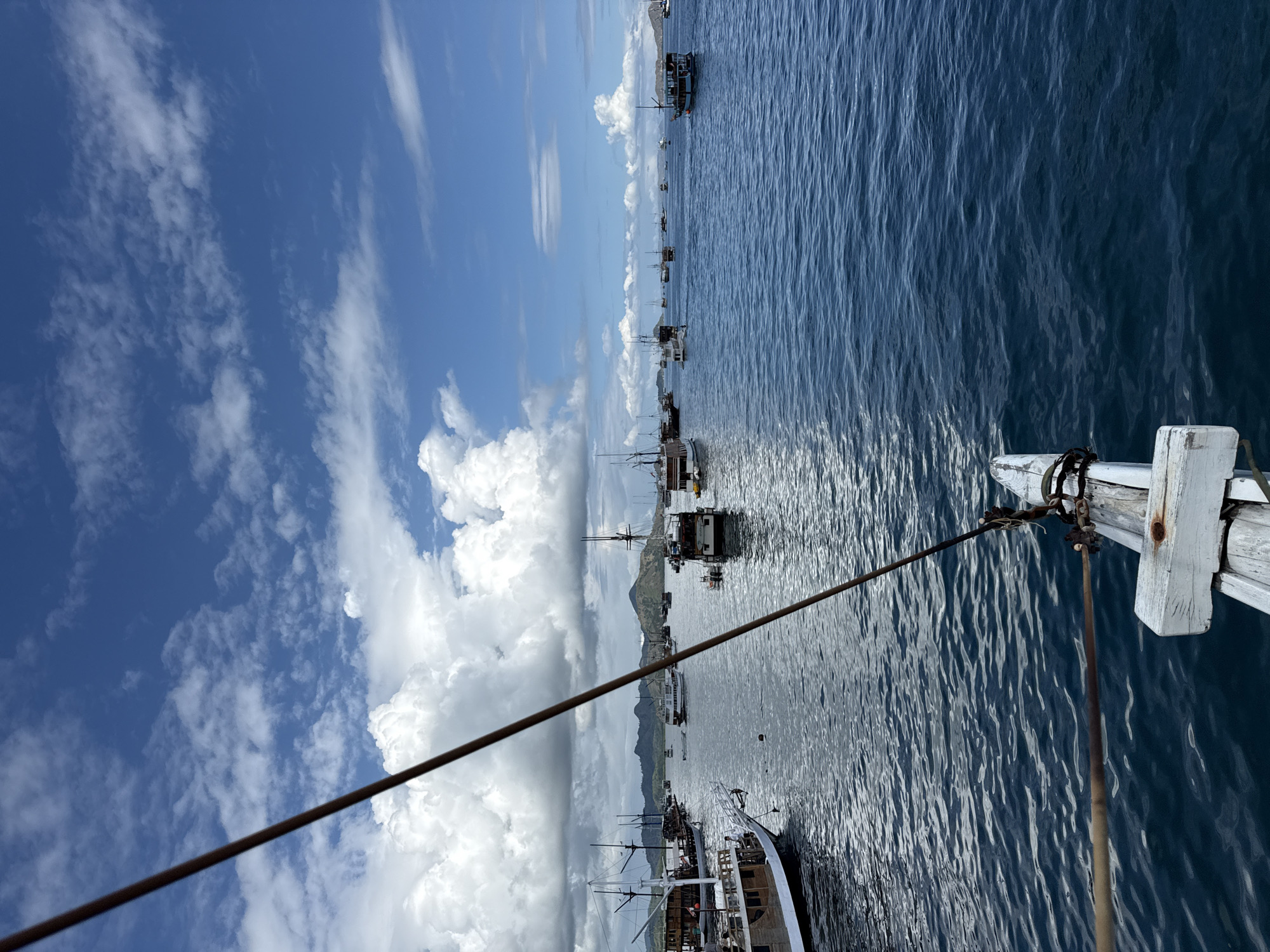 The image is taken from a boat in Labuan Bajo, Indonesia, looking towards the sea and coastline. Numerous boats are visible in the water, ranging from small vessels to larger ships. A land mass or island is visible in the distance, partially obscured by clouds. The sky is blue with puffy white clouds. A rope stretches from the bottom left to the upper right of the frame, intersecting with part of the white wooden structure of the boat the photo is taken from. The sea has a dark blue color with ripples and light reflections.