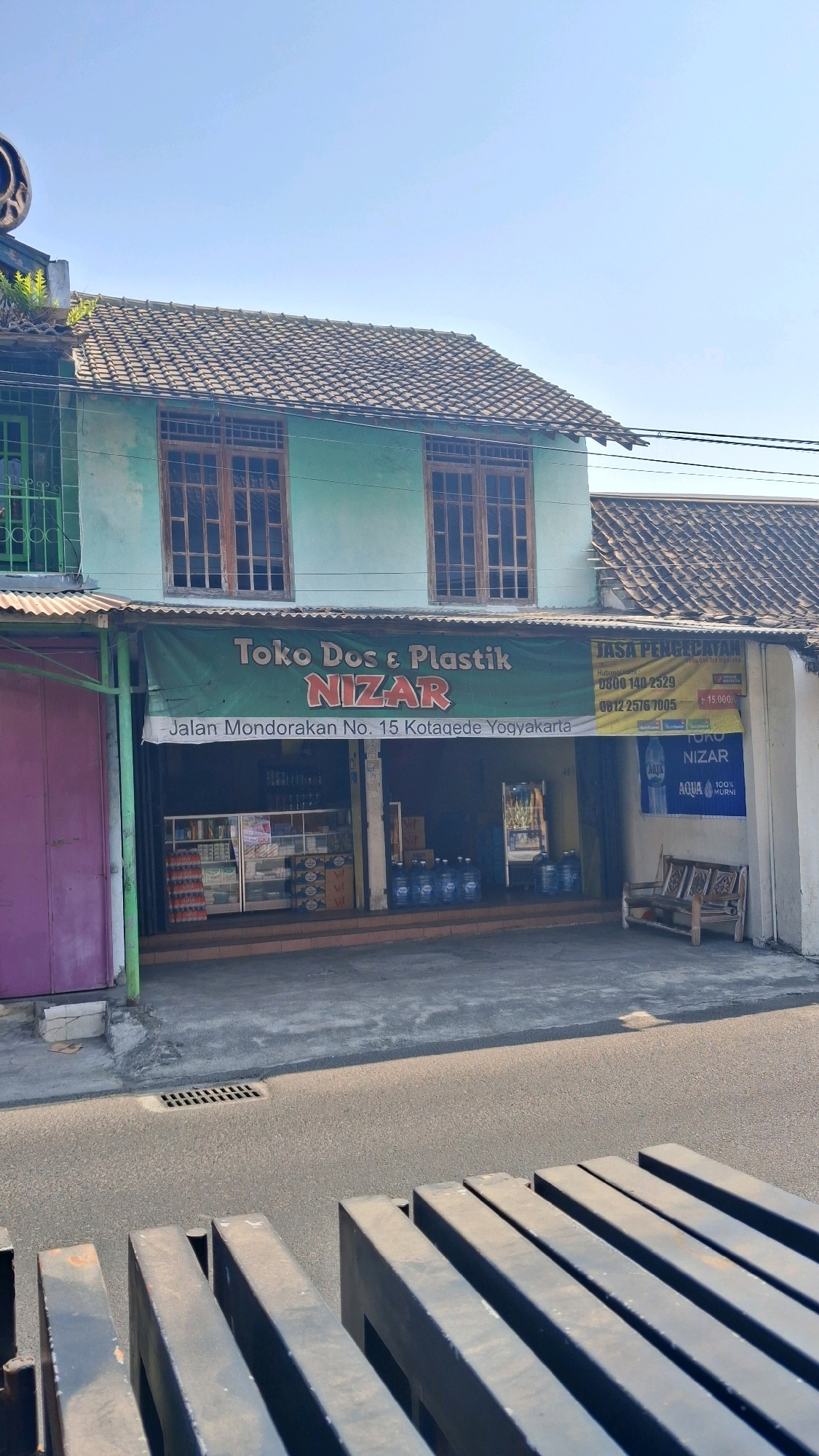 The image shows a storefront in Banguntapan, Indonesia. The storefront is for "Toko Dos & Plastik NIZAR" and is located on Jalan Mondorakan No. 15 Kotagede Yogyakarta. The building has a light green facade with wooden-framed windows. A green banner above the storefront displays the store's name and address. Inside the store, shelves are stocked with various goods, including bottled water and other products. In front of the store, there are several large blue water jugs. A wooden bench sits to the right of the entrance. The street in front of the store is paved, and there is a metal grate in the road. The sky is clear and blue.
