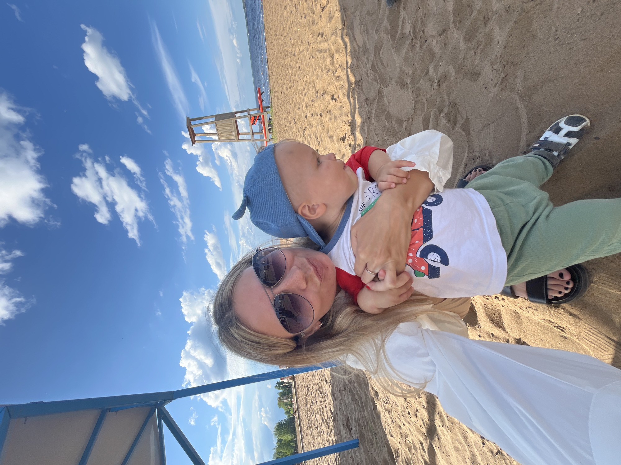 This media file captures a mother and her young child enjoying a sunny day at a sandy beach in Chaikovsky, Russia. The subjects are a woman and a baby. The woman, wearing large sunglasses and a white top with voluminous sleeves, is embracing the child, who is dressed in a blue cap and a white t-shirt with a colorful graphic.
The scene is a beach with fine, light-brown sand. In the background, a calm body of water, likely a river or lake, is visible, meeting the horizon under a bright blue sky dotted with fluffy white clouds. A lifeguard tower stands on the sand near the water's edge, suggesting this is a public beach. Part of a blue and beige beach canopy is visible in the foreground.
The image appears to have been taken during daylight hours, with ample sunlight casting clear shadows and illuminating the scene. The overall mood seems relaxed and happy, indicative of a pleasant family outing. The close embrace between the mother and child suggests affection and a sense of contentment.
There is no visible text in the image.