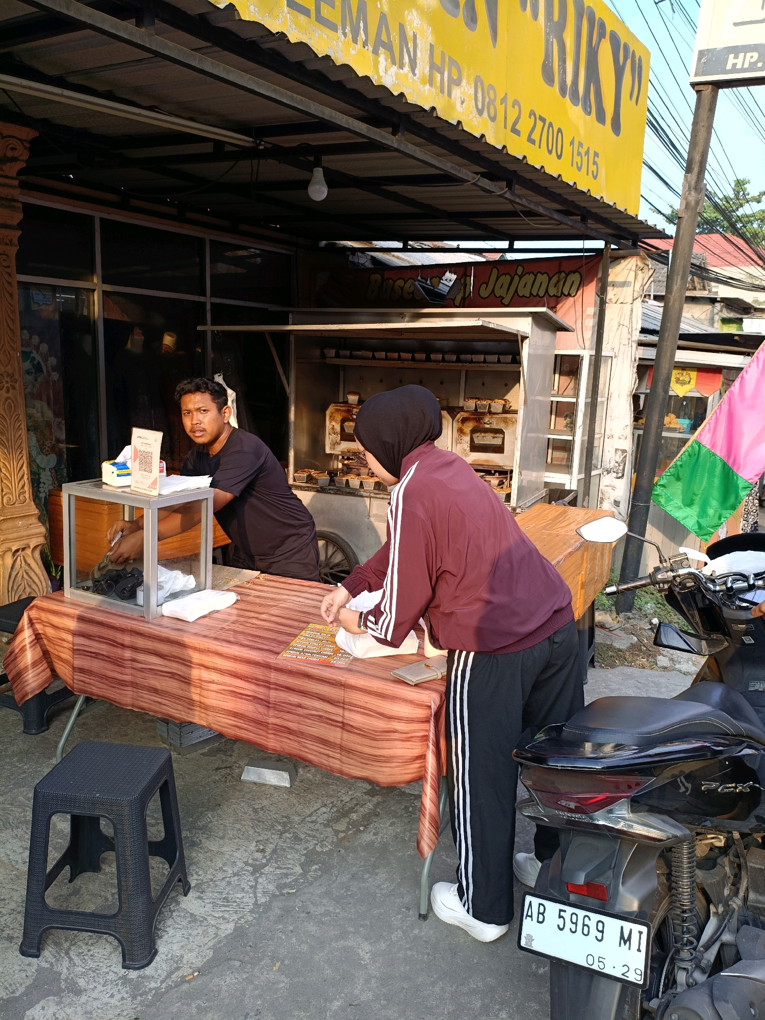 In Maguwoharjo, Indonesia, on a sunny day, a street food stall is shown with a man and a woman attending to customers. The stall features a yellow awning with text including "RIKY" and a phone number, under which a menu is displayed. A woman in a black hijab and a maroon jacket is interacting with the food stall. A man, possibly the vendor, is wearing a black t-shirt and looking towards the camera while attending to a customer. Behind them, a food cart with baking trays is visible. A black scooter is parked to the right of the stall, with a license plate reading "AB 5969 MI". The ground is paved and there are power lines overhead.