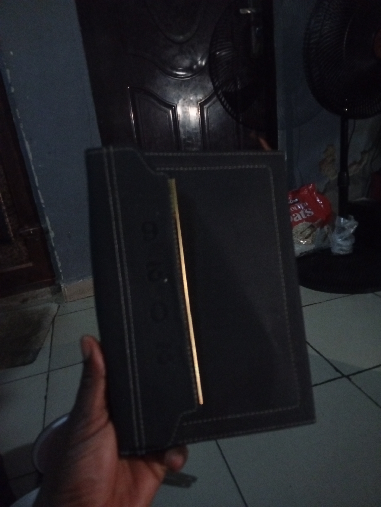 An over-the-shoulder, low-angle shot, taken indoors, shows a dark-skinned hand holding a black diary. The diary has a gold strip running vertically down the center of its front cover. The diary also has the embossed numbers "9", "2", and "0" visible down its left side. The background is dimly lit and shows a dark door, a standing fan, and a bag of oats. The floor is tiled. The image does not provide any information to determine the location in the city of Ashata, Nigeria.