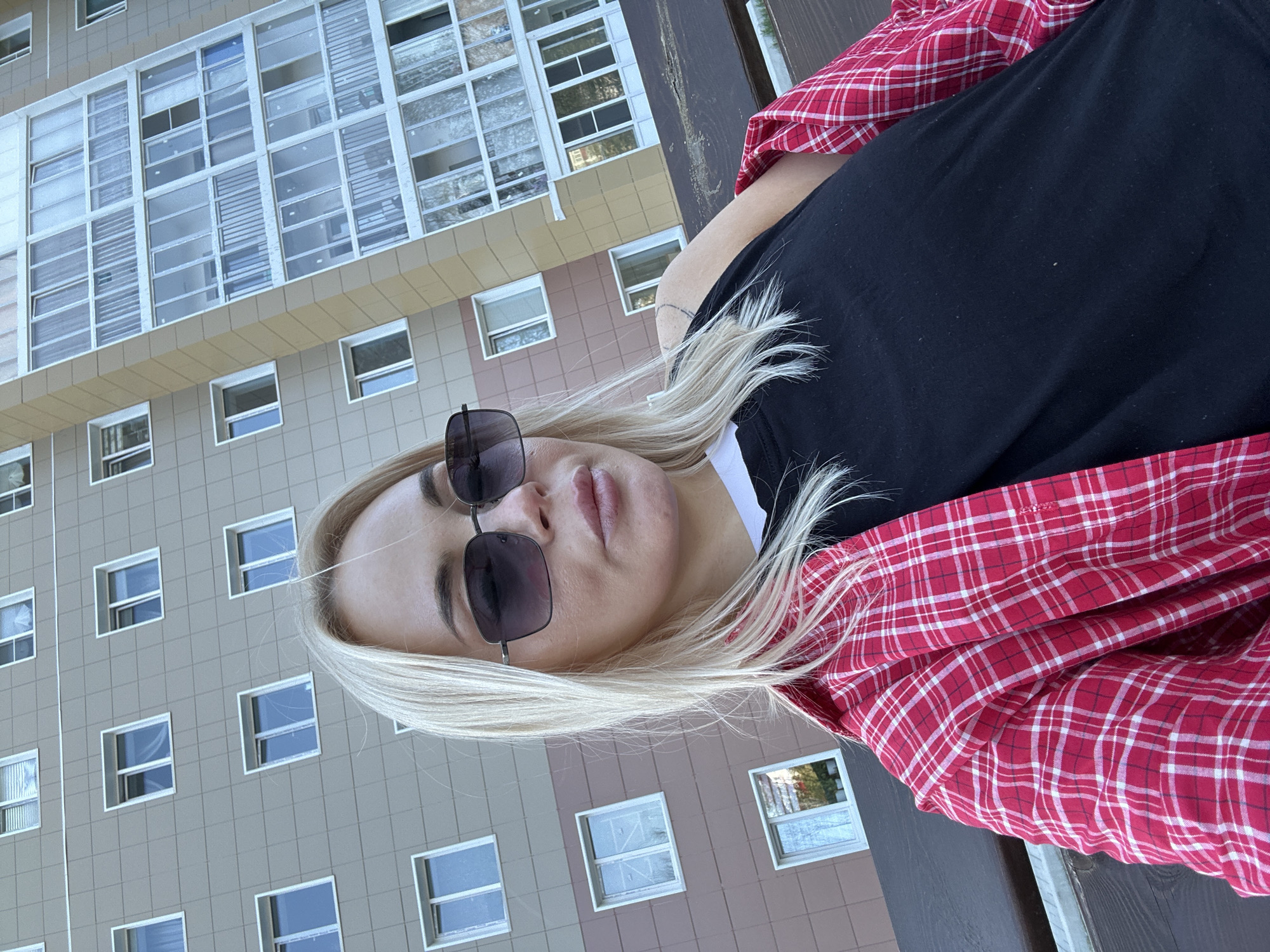 A fair-skinned woman with blonde hair, wearing sunglasses, a black shirt, and a red-and-white plaid shirt, is oriented in a portrait-style composition against a background of an apartment building. The building, located in Megion, Russia, is characterized by repetitive rectangular windows set within a light-colored facade.