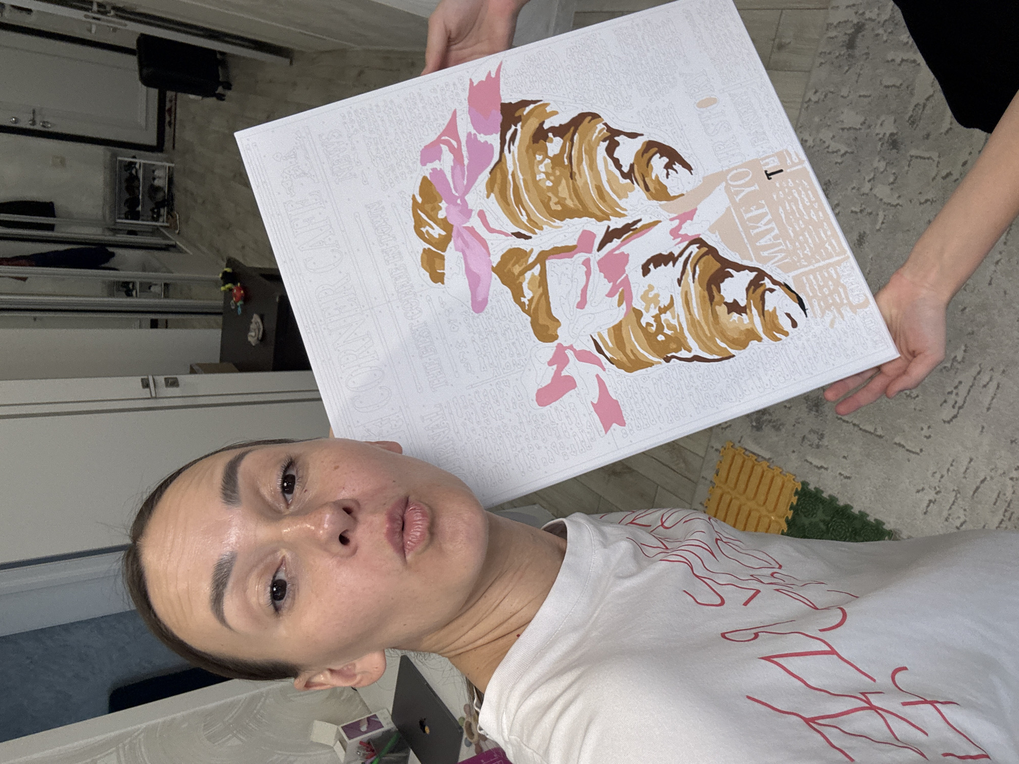 An indoor selfie photograph depicts a woman posing in what is identified as Sochi, Russia. The woman, appearing to be in her mid-to-late 30s, is positioned in the left foreground, looking directly at the camera. Her light brown hair is pulled back, revealing a high forehead. She has brown eyes, and visible freckles dot her nose and upper cheeks. Her lips are pursed in a "kiss" or "pout" expression. She wears a white short-sleeved t-shirt adorned with red abstract, flame-like designs on the front. Her right arm is extended, holding the camera device (unseen).
The primary subject of the photo, besides the woman, is a rectangular white canvas, held vertically in the lower-right portion of the frame. This canvas is a "paint by numbers" project, featuring a pre-printed design of two croissants, partially painted in shades of brown and tan. Pink bows are depicted on and around the croissants, some also filled with pink paint. The canvas background displays faint outlines of text, resembling newspaper print, with phrases such as 'CORNER CAFE,' 'NEWS,' 'THE BEST COFFEE IN TOWN,' and 'MAKE YOUR STORY.' Numerous small, numbered sections remain unpainted. The hands and forearms of a second individual, wearing a black long-sleeved garment, are visible at the bottom right, assisting in holding the canvas.
The background reveals an interior setting, likely a hallway or entrance area within an apartment or house. The flooring consists of light-colored laminate, possibly wood-imitation. A grey patterned rug is visible beneath the canvas and extending to the left, and yellow and green interlocking foam floor mats are seen on the far left, suggesting a children's play or exercise area. Two white doors with silver-colored handles are visible on the right, and another partial white door is on the far left. A large mirrored wardrobe occupies the left background, reflecting interior shelves with various garments (blue and red) and shoes. A small white metal shoe rack, laden with multiple pairs of shoes (primarily white sneakers), stands against the wall between the mirrored wardrobe and the right-side doors. A dark brown, low rectangular table with some small objects rests near the bottom left. A black hard-shell suitcase on wheels is positioned near the far right door. A desk or table with a dark laptop (featuring an Apple logo) and a purple container with pens is partially visible on the far left. The scene is illuminated by ambient indoor lighting.