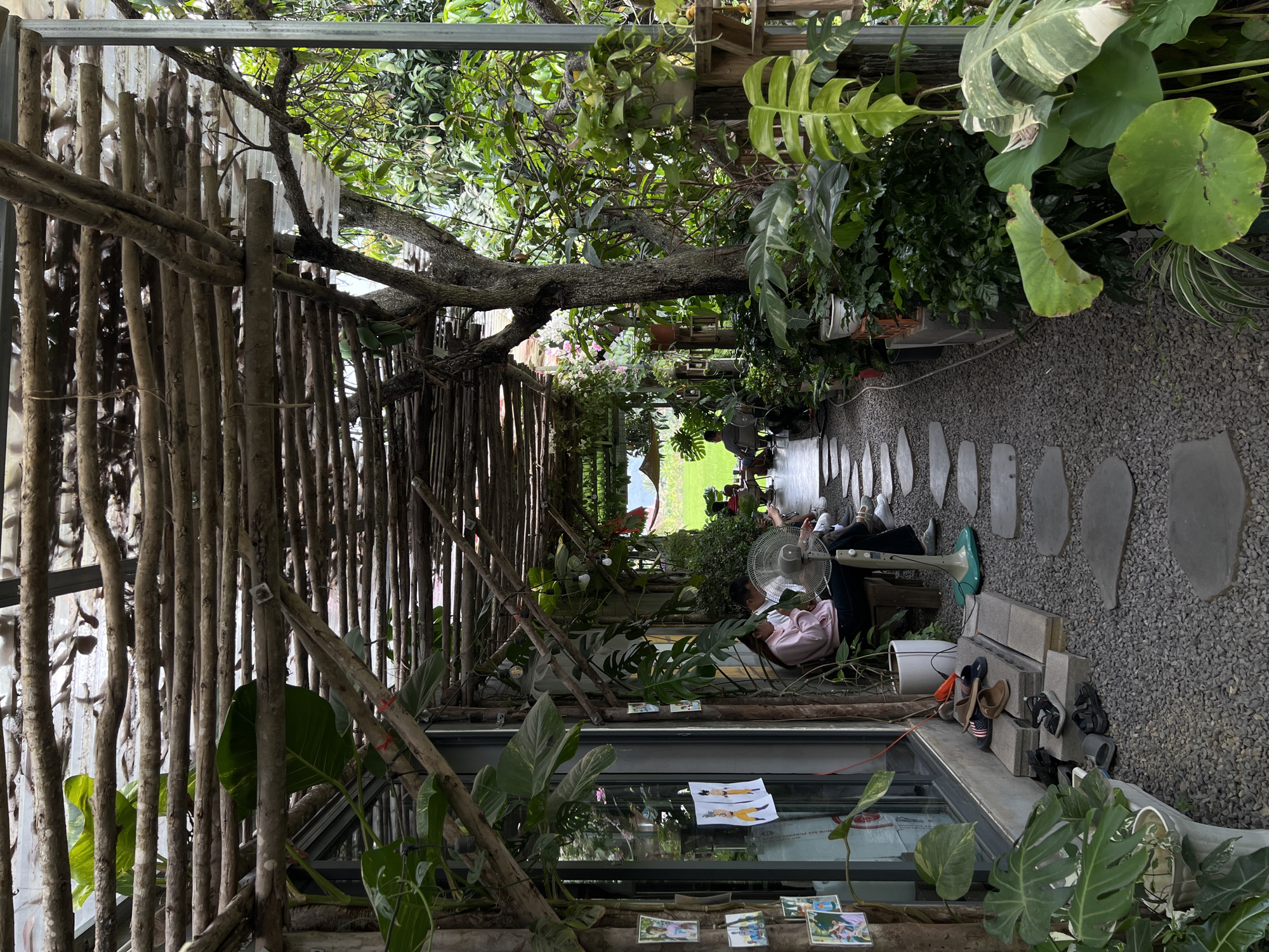 A gravel walkway with stone pavers is flanked by lush greenery. A rough wooden structure covers the path. A person sits near a fan. Shoes are arranged near a small ledge. A building sits on the right. Decorations are attached to the building. The image appears to be in Diên Khánh, Vietnam.