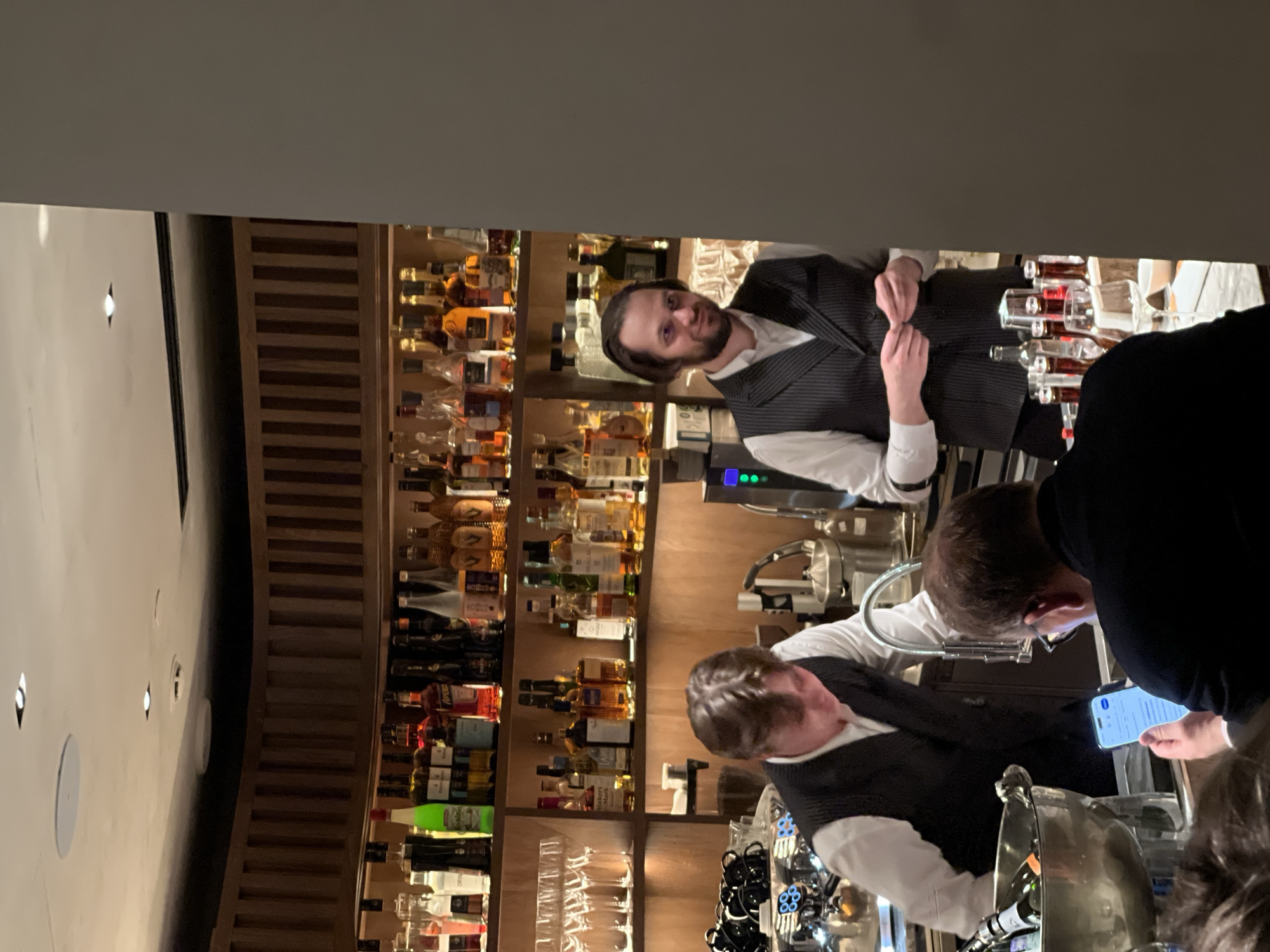 The image shows a bar in what appears to be a high-end establishment. Behind the bar, there's a large display of liquor bottles arranged on shelves. Two bartenders wearing vests and white shirts are behind the counter. One bartender is facing the camera, while the other seems to be interacting with a customer who is looking at a phone. There is a wine chiller in front of the counter, with a bottle inside. I have no information to assume the bar is located in Moscow, Russia.