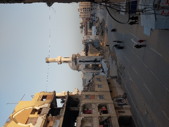 The image captures a street scene in a heavily damaged urban environment, likely following a conflict or disaster. The perspective is elevated, looking down onto a paved road flanked by severely damaged buildings.
On the left side of the frame, occupying the foreground and mid-ground, are multi-story buildings that have suffered extensive structural damage. Facades are partially collapsed, revealing the interiors of rooms filled with rubble and debris. Exposed rebar and broken concrete are visible, indicating significant destruction. One building closest to the viewer has a large section of its wall missing, exposing multiple floors.
In the center-left, standing amidst the destruction, is a mosque with a tall, slender minaret and a dome. The mosque itself appears damaged, with visible signs of structural compromise, particularly around its base and dome area.
The paved street, running from the mid-ground towards the background on the right side of the image, has clear white lane markings and a white arrow painted on the surface, indicating direction. The road surface appears dusty or covered with light debris. On the right side of the street, there are several white tent-like structures, possibly temporary shelters, set up amidst the rubble.
Several people are visible walking on the street. In the mid-ground, a group of approximately 4-5 individuals, mostly dressed in dark clothing, are walking on the right side of the road, moving generally towards the viewer. Further down the street, additional smaller groups or individuals can be seen. Their expressions and specific details are not discernible due to the distance.
The background shows more urban buildings, some appearing less damaged than those in the foreground, extending towards the horizon. The sky is clear and light blue, suggesting daytime, possibly late afternoon or early morning due to the soft light, though no strong shadows are cast.
The overall impression is one of widespread devastation and an ongoing sense of daily life continuing amidst significant ruin. No discernible text is visible within the image. A distinct vertical white line with dark edges runs through the image, particularly noticeable in the sky and across the minaret, which appears to be an image artifact rather than a part of the scene.