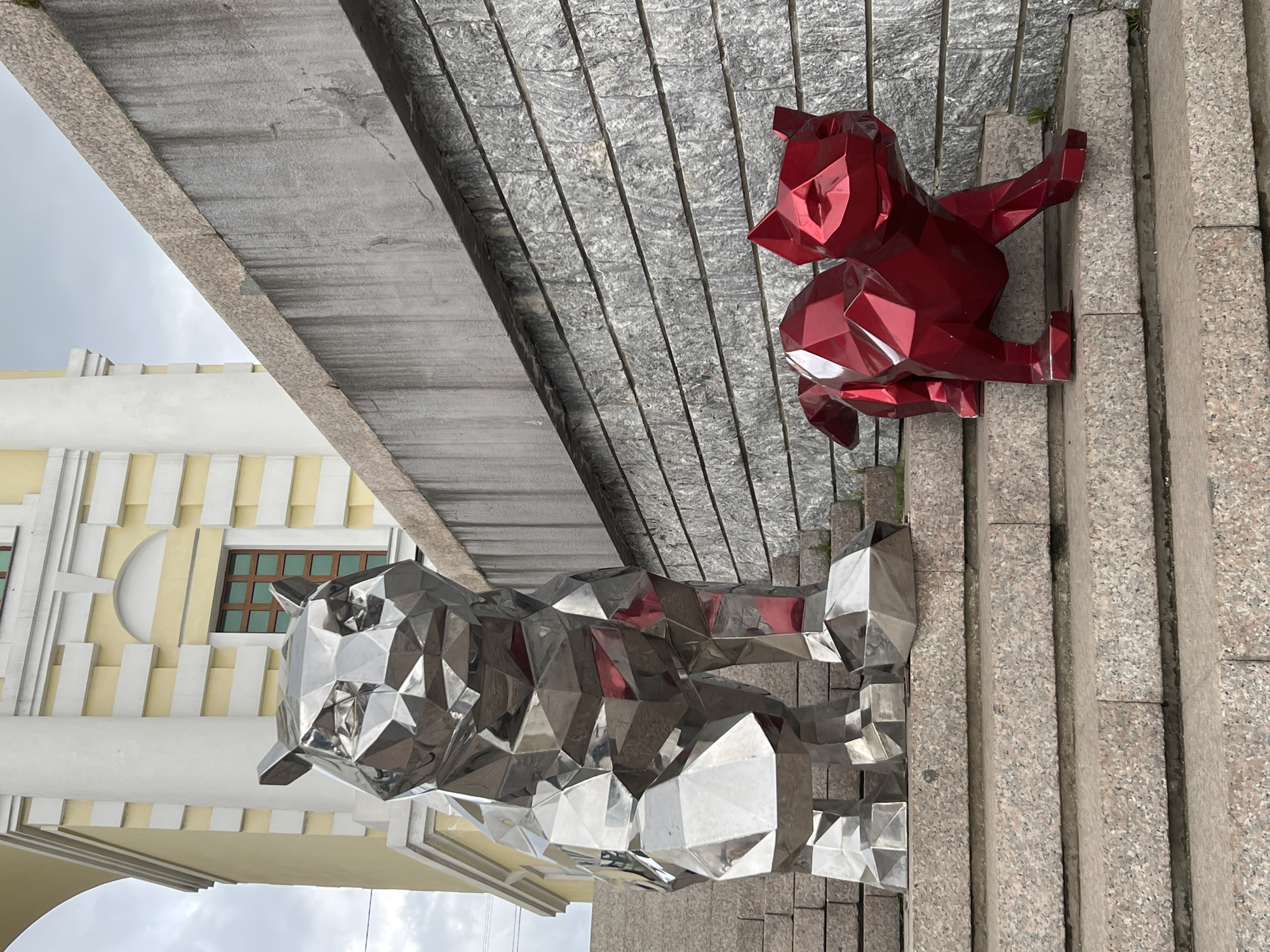 The image shows two polygonal animal sculptures, a silver bear and a red wolf, located in Irkutsk, Russia. The silver bear is standing while the red wolf is sitting, positioned on stone paving and steps. Part of a yellow and white building with a window can be seen in the background, under an overcast sky. The scene appears to be an urban public space.