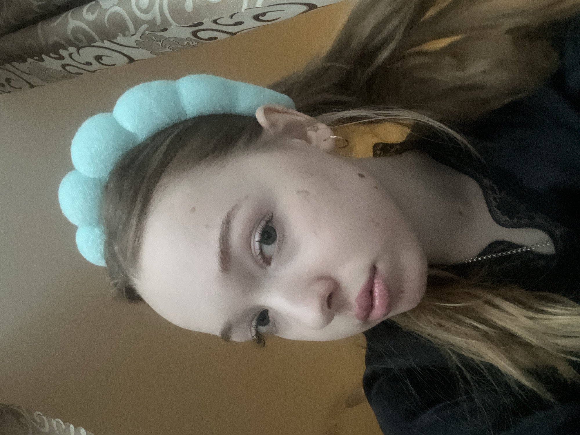 A young woman poses for a selfie indoors. She is wearing a light blue, cloud-shaped headband. Her hair is blonde. She is wearing a black top with lace detail and a silver necklace. She appears to be inside a residence. The location in the city of Chaikovsky, Russia is not evident.