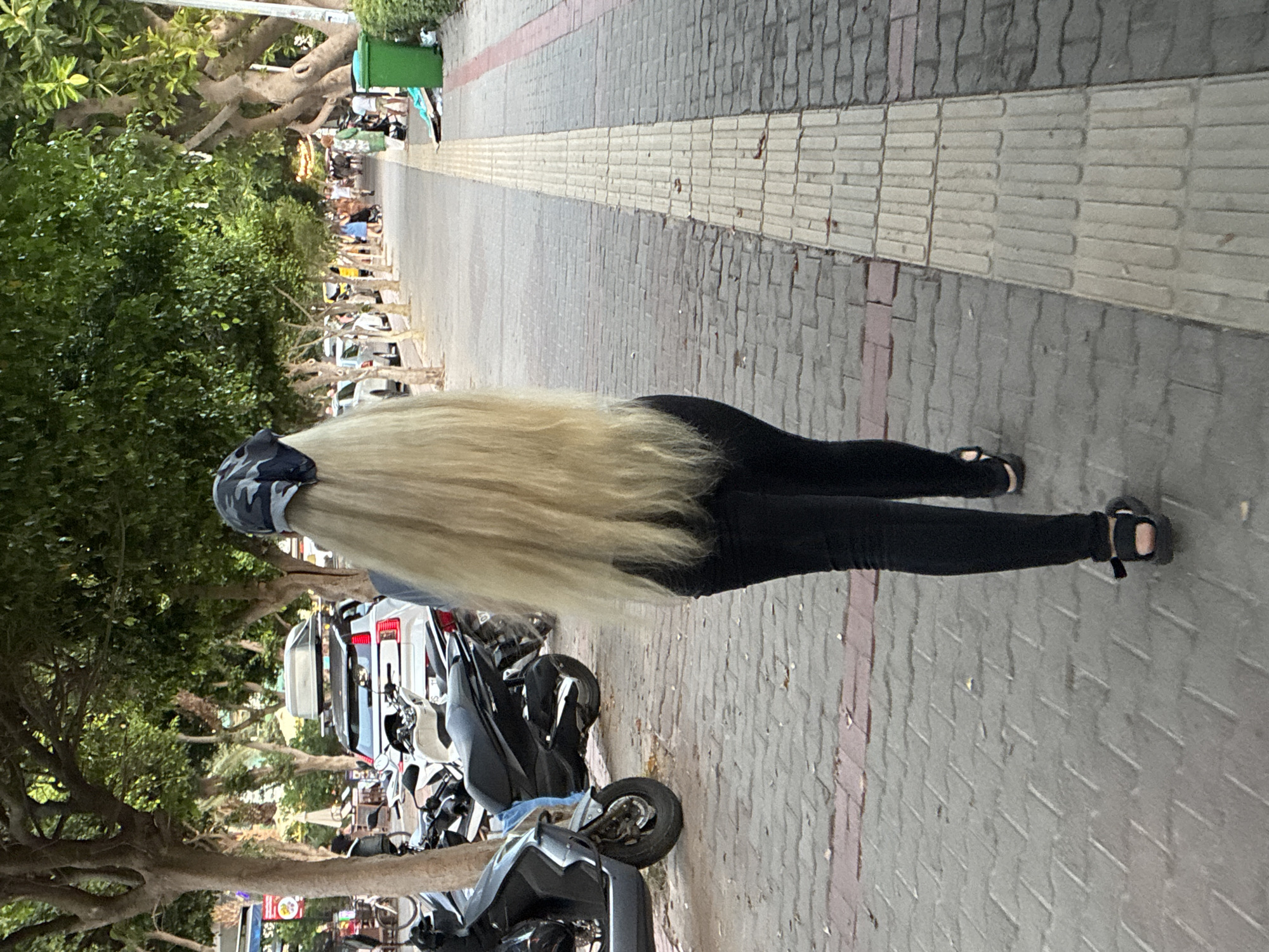 A person with long, blonde hair and a camouflage bandana walks along a paved sidewalk lined with trees in Alanya, Turkey. The person is wearing black pants and black sandals. To the left of the sidewalk, several scooters are parked, and further back, a white SUV with a roof carrier is visible. The sidewalk is bordered by a strip of tactile paving. In the background, other pedestrians are walking along the sidewalk, and buildings and more trees are visible. A green trash can stands on the right side of the walkway. The scene appears to be a street or promenade in a city.
