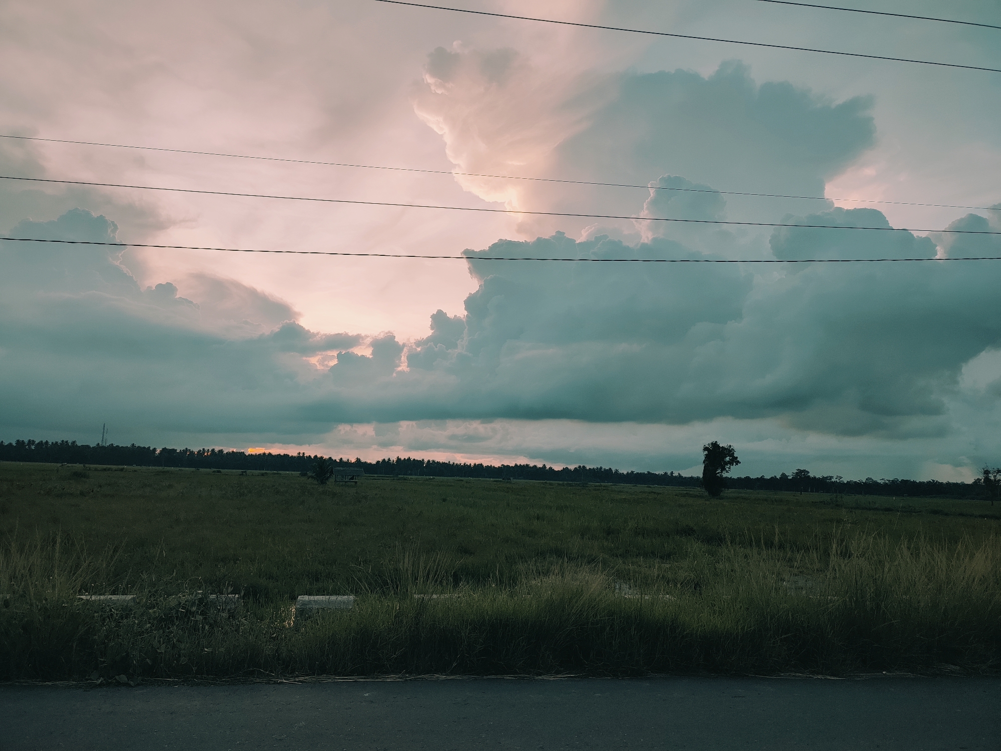The image depicts an open field scene in Muliengmanyang, Indonesia. The foreground includes a paved road with overgrown vegetation between the road and the field. The field itself is grassy and extends towards a line of trees on the horizon, behind which a sliver of sunset-colored sky is visible. Above, a dramatic sky is filled with large, cumulus clouds, some with a pinkish hue. Electrical wires stretch across the upper portion of the frame.