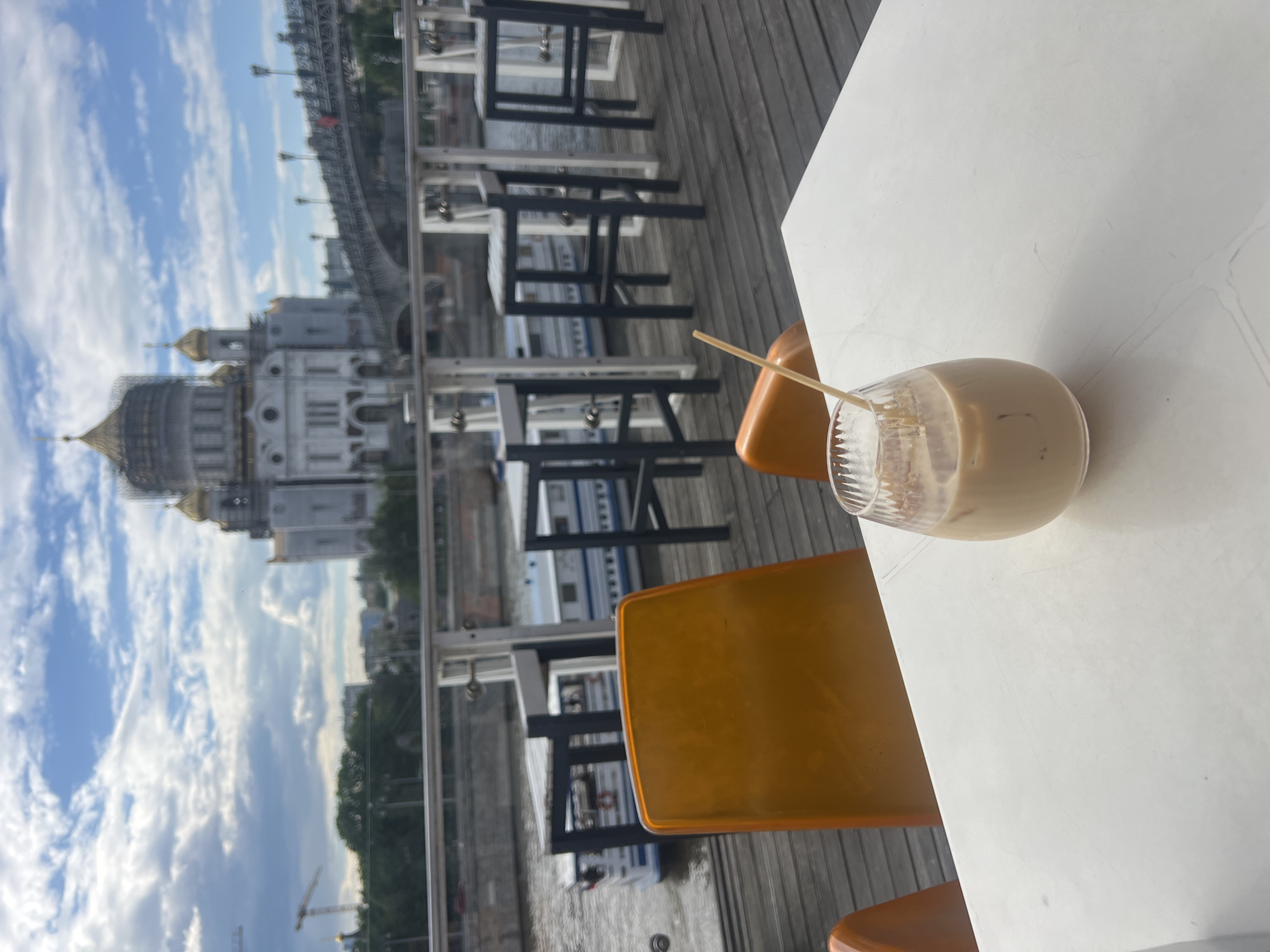 A glass of iced coffee with a straw sits on a white table, with orange chairs in the background. Through the glass railing, the Cathedral of Christ the Savior and a bridge are visible in the city of Moscow, Russia. Other tables, chairs, and boats can be seen along the river. The sky is cloudy.