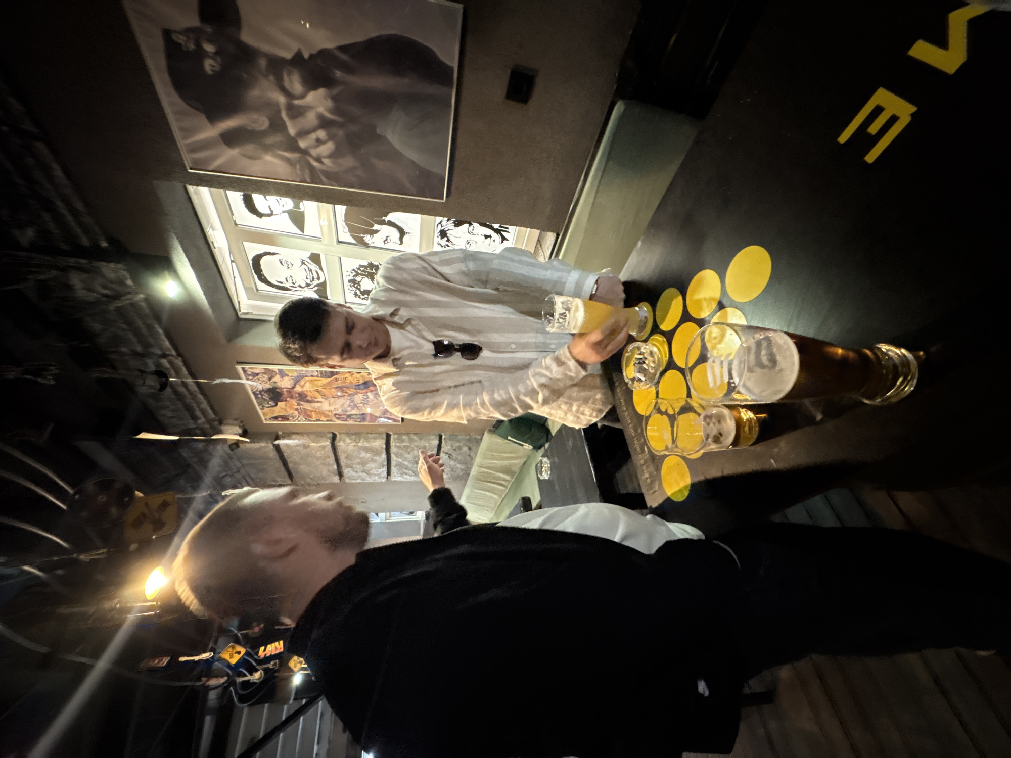 A medium shot shows two people inside a pub in Minsk, Belarus. One man wears a striped shirt and is pouring beer from one glass to another. Sunglasses rest in front of him on the dark table, which has yellow circles painted on it. The second person is on the left, and the shot focuses on the top of his head. He wears a black shirt. In the background are many photos on the wall and hanging from the ceiling.